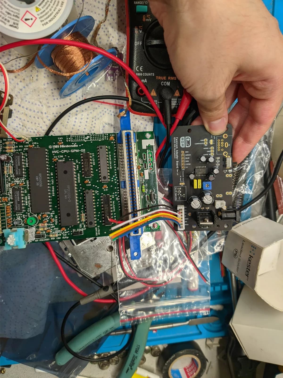 A hand holding the small black composite mod board beside the green Nintendo Famicom motherboard (CPU-GPM-02) on the workbench, with a multimeter and bagged replacement parts sitting nearby.