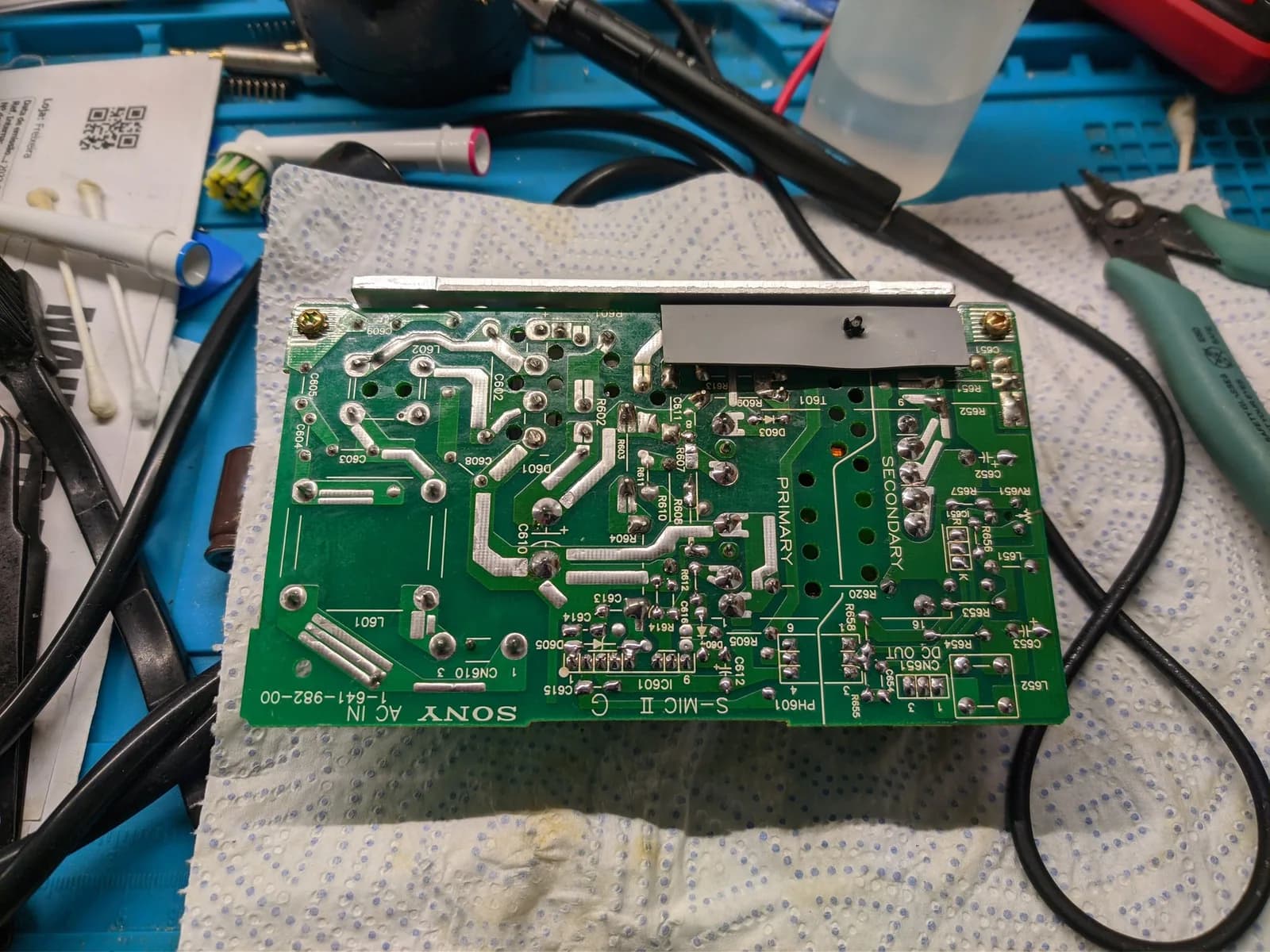 Sony BVM-9045QD PSU board recapped, showing green PCB with replaced capacitors, soldering iron, and work-in-progress restoration on blue mat.