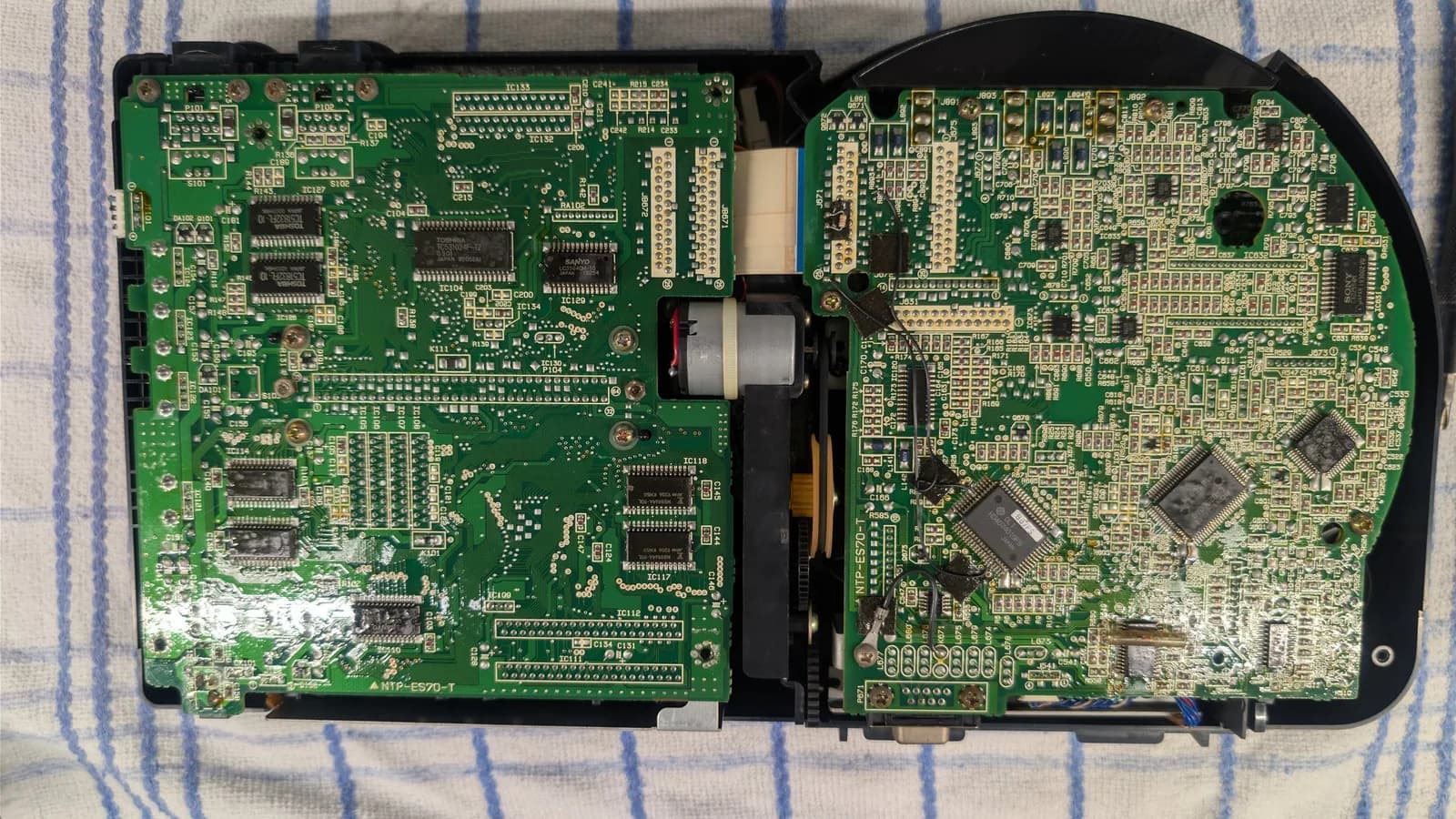 Sega Wondermega HWM-5010 opened for disassembly, showing two large interconnected circuit boards packed with ICs and connectors, with the grey CD drive motor, belt and gears in the gap between them.