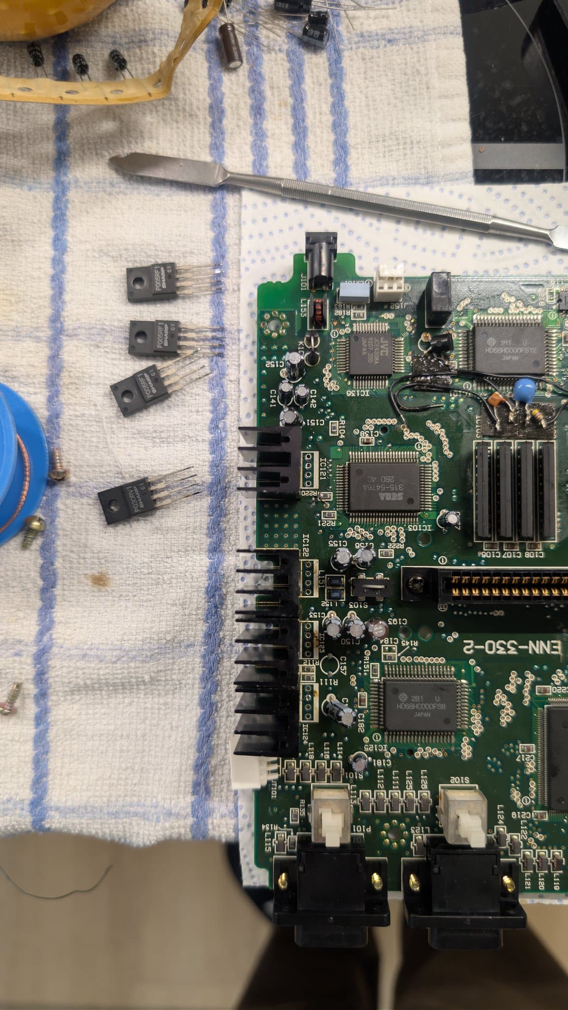 Sega Wondermega HWM-5010 main board with five removed voltage regulators on cloth, tweezers, and capacitors visible during recapping and regulator cleaning repair.