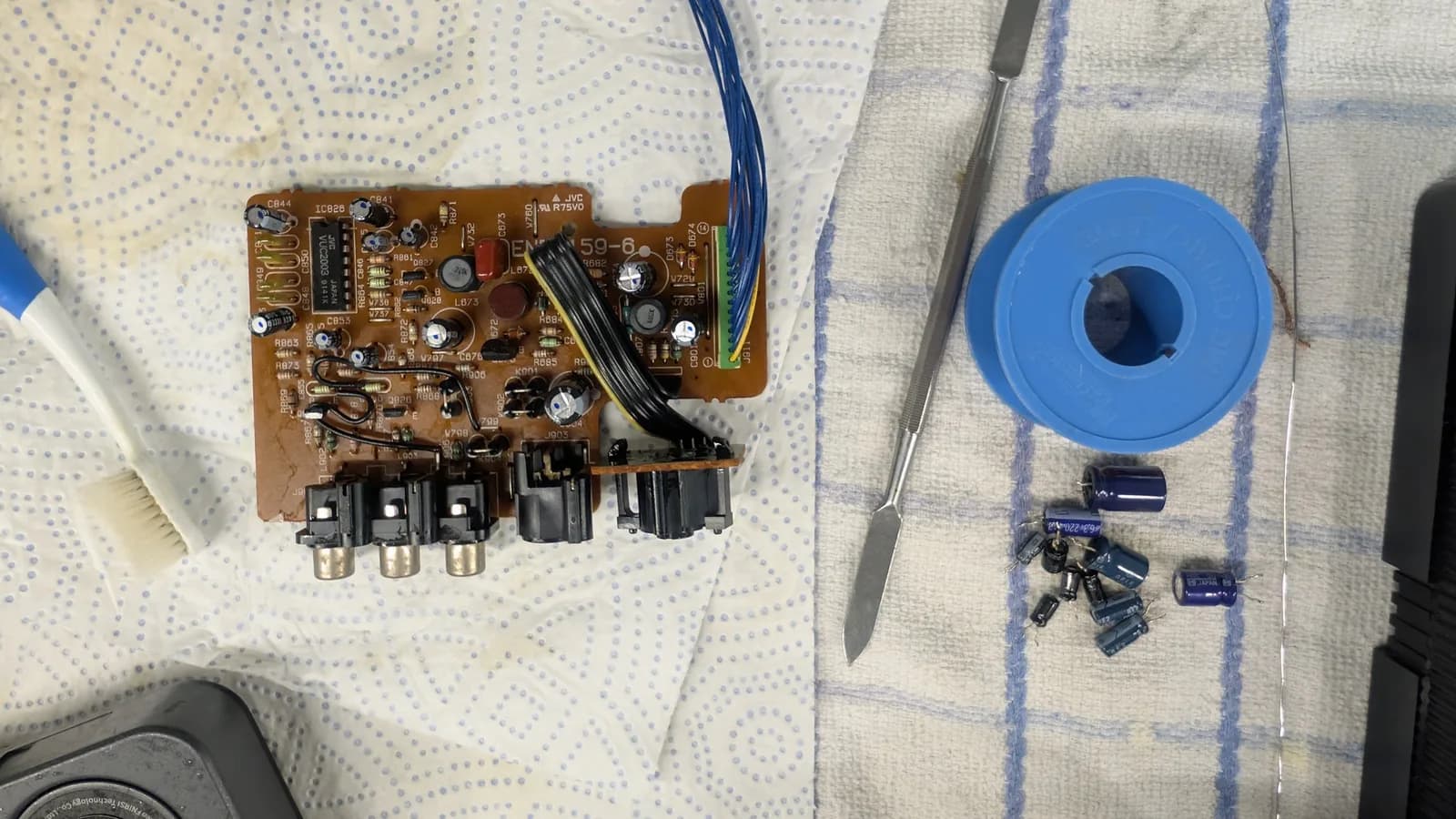Brown phenolic Sega Wondermega video board (JVC VUC2003 IC, RCA output jacks) recapped, its new capacitors marked with a blue dot, with a cleaning brush, a metal spatula, a blue spool of solder/braid, and the removed old capacitors piled on the towel.