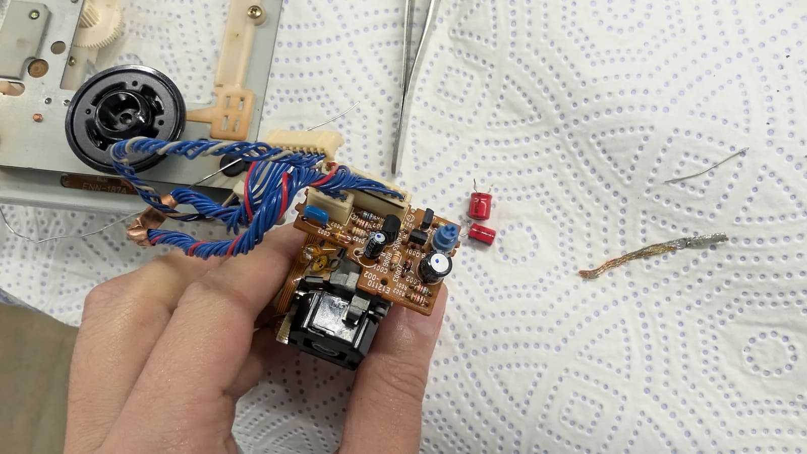 Recapped laser pickup PCB (silk-screen E12110-002) held in hand, its new capacitors marked with a blue dot and the old red capacitors on the paper towel below, with blue and red twisted wire pairs and the JVC Optima-5 optical pickup block and objective lens at its base.