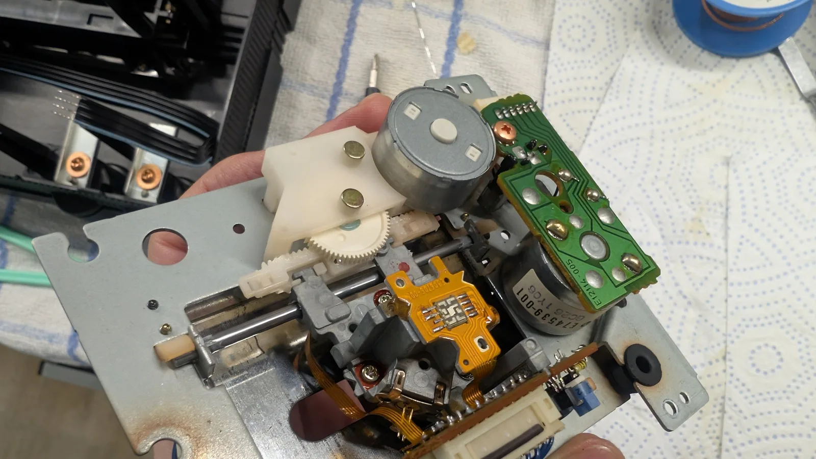 CD laser traverse assembly held in hand, cleaned and with its moving parts lubricated with Molykote DX paste, showing white plastic gears, the worm-drive shaft, the sled motor, an orange sensor flex, and a small green control PCB (silk E12114-005).