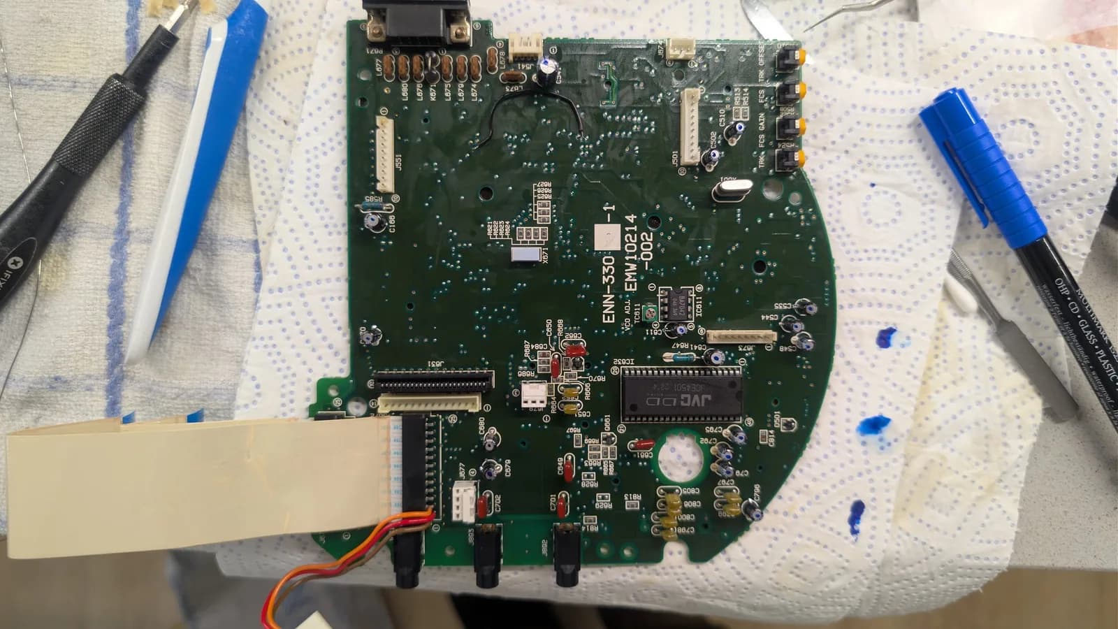 Sega Wondermega CD-ROM/servo board (silk-screen ENN-330-1, JVC servo IC, CD adjustment points) fully recapped, its new capacitors marked with a blue dot, with an iFixit driver, a pry tool, tweezers, and the blue marker pen on the towel.