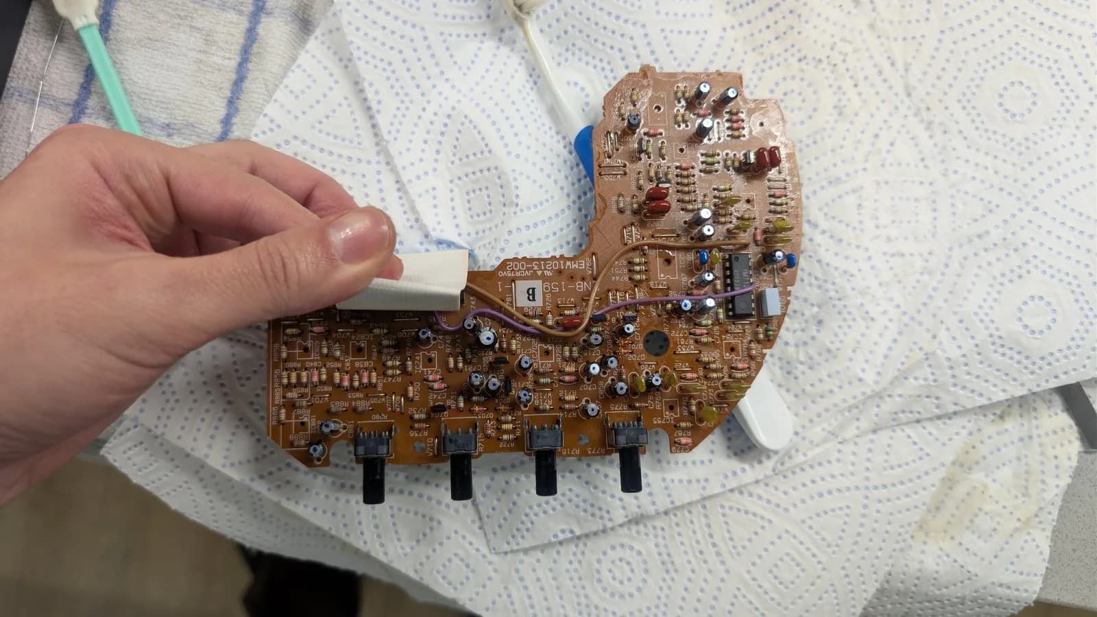 Hand holding the recapped Sega Wondermega sound/control board, its new capacitors marked with a blue dot, showing four black potentiometers with knurled shafts along one edge.