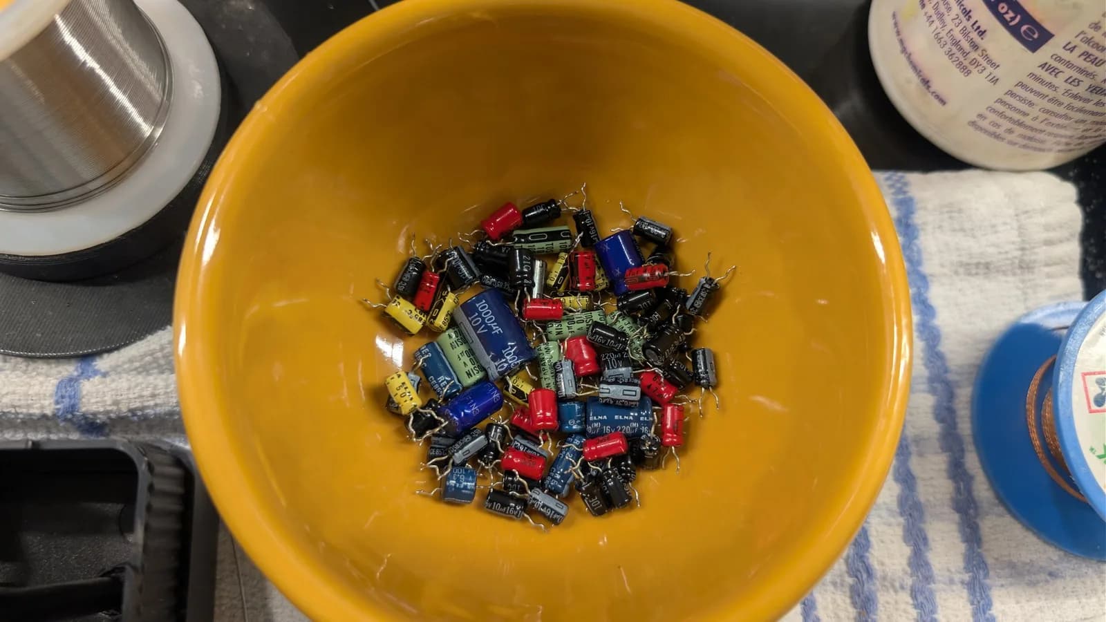 Yellow bowl holding dozens of desoldered electrolytic capacitors (ELNA and others) removed from the Sega Wondermega HWM-5010, with a solder spool and an MG Chemicals bottle behind it.