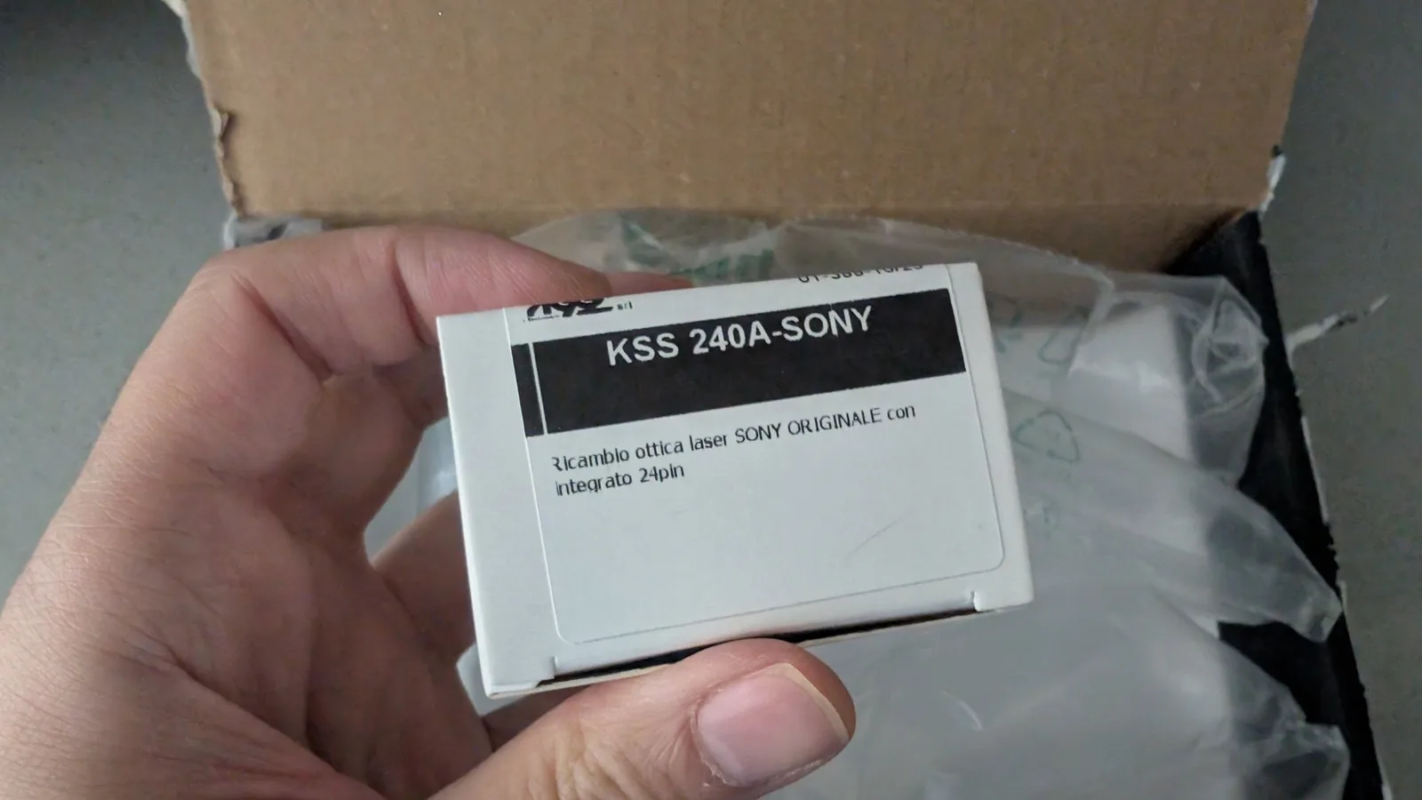 Hand holding KSS 240A-SONY laser pickup with packaging in cardboard box, replacement optical component for Mega-CD repair.