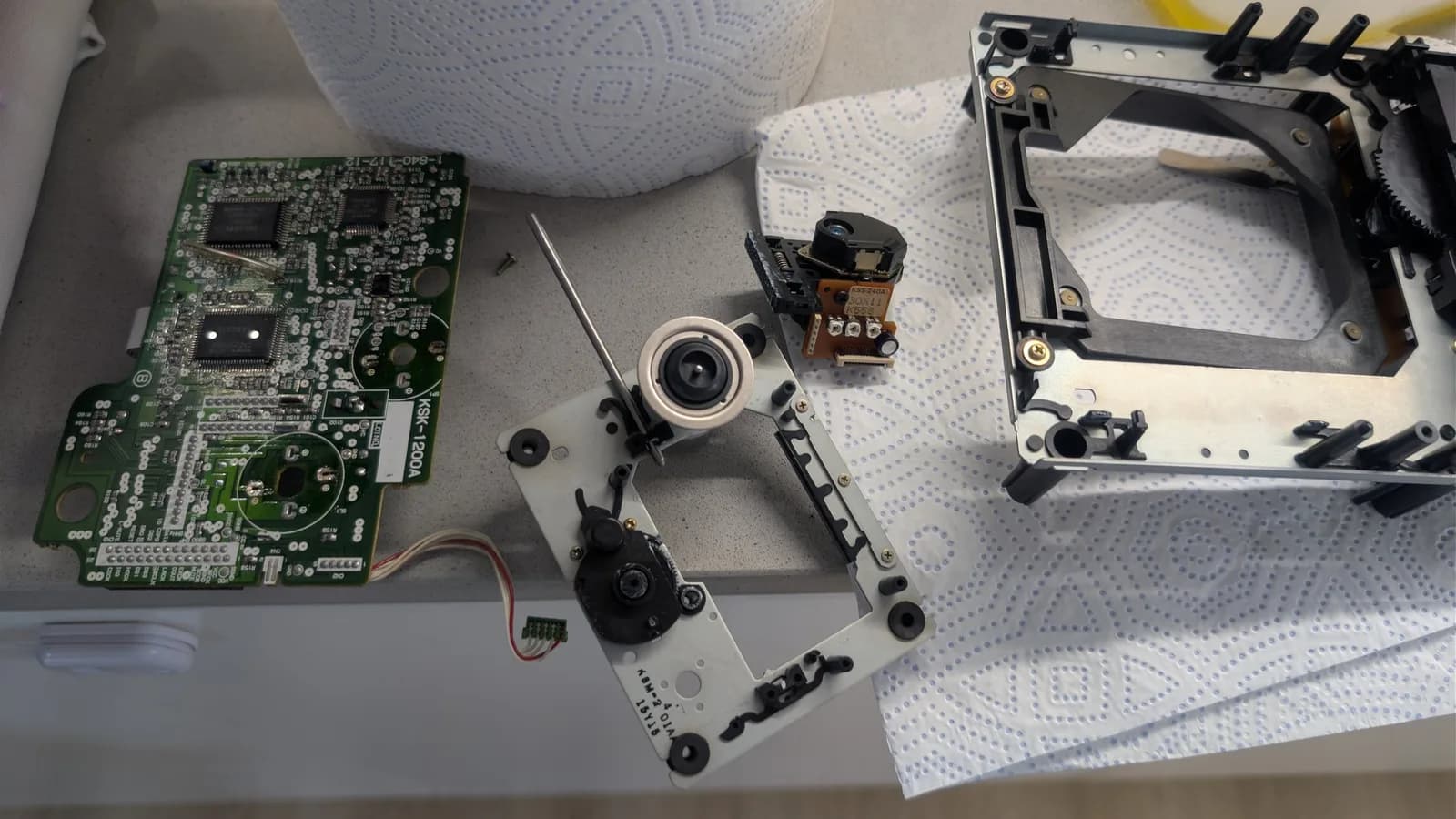 Disassembled Mega-CD 1 showing green mainboard, KSS-240A optical pickup, motor assembly, and metal chassis components laid out on work surface.