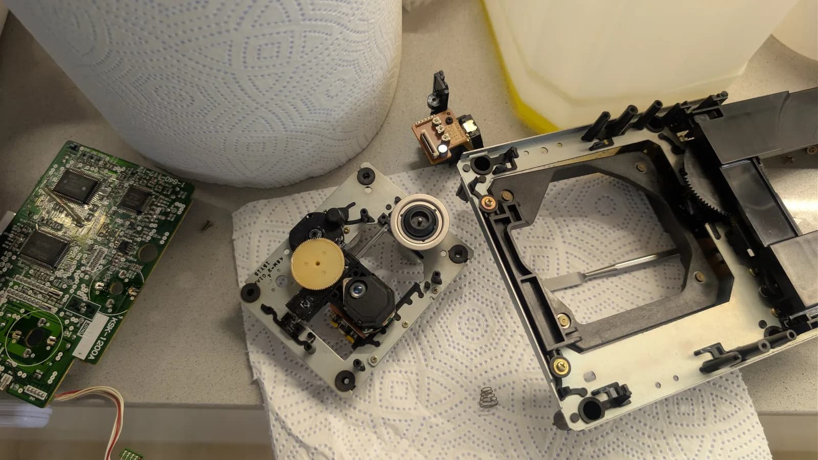 Disassembled Mega-CD 1 optical drive showing new laser pickup installed, circuit board, and mechanical housing components on work surface.