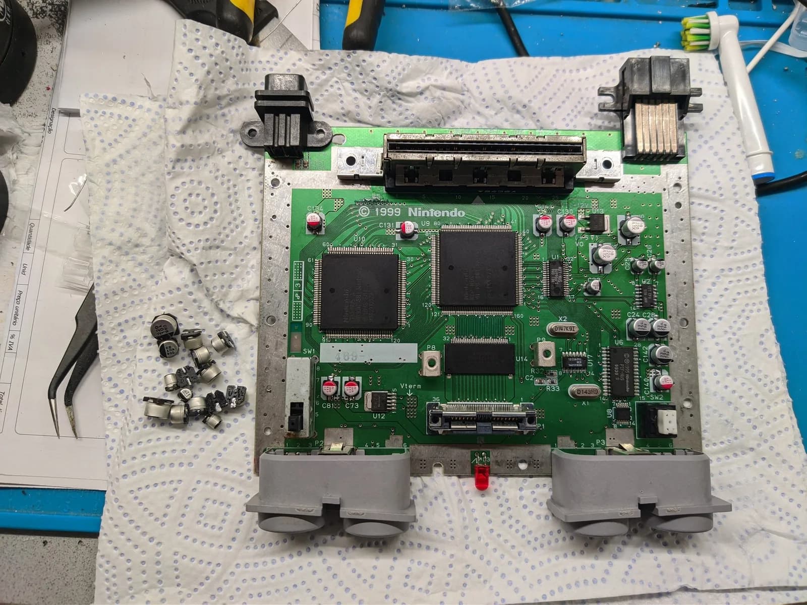 Nintendo 64 motherboard after recapping, the new SMD electrolytic capacitors (grey metal cans) already installed on the board, with the old ones removed and set to the left, on a paper towel over a blue silicone mat.