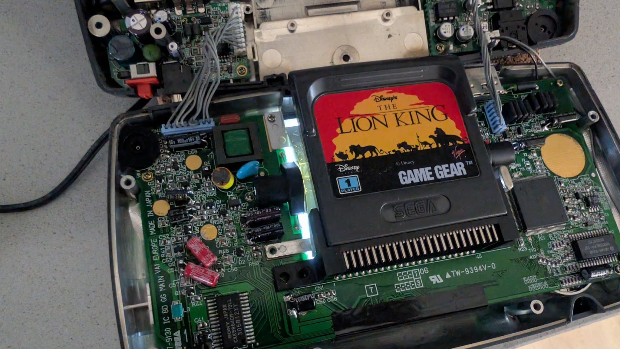 Sega Game Gear interior with cartridge slot open, showing motherboard, CCFL backlight assembly, and circuit boards during repair inspection.