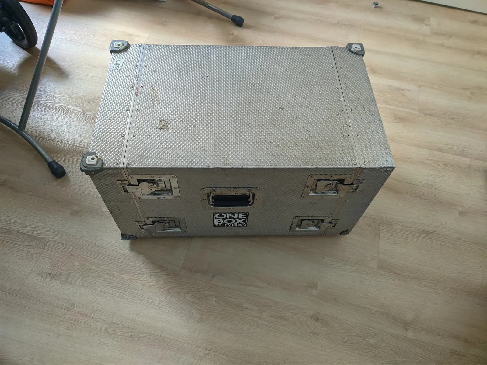 Closed silver aluminium flight case with hard plastic corner protectors and 'ONE BOX TELEVISION' branding, on a wooden floor.