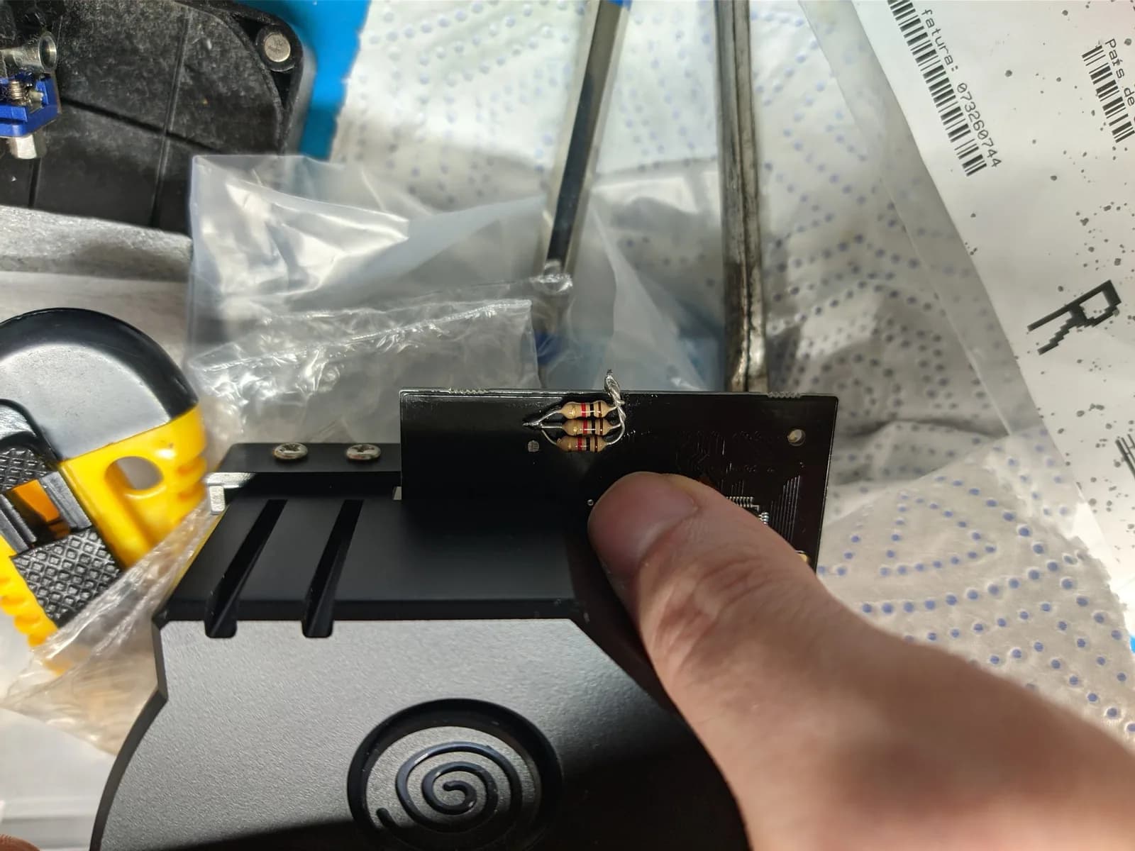The underside of the black GDEMU PCB resting on a paper towel over a blue silicone mat, showing the three axial resistors of the follow-up 12V-line heat mod, with one end of each resistor soldered to a point on the board's underside.