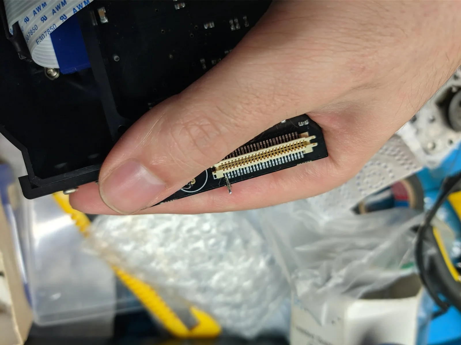 The top side of the black GDEMU PCB, its fine-pitch gold-pin connector held between finger and thumb, showing a metal pin soldered onto one connector pin as the tie-point where the other end of the 12V-line heat resistors attaches; the workbench behind.