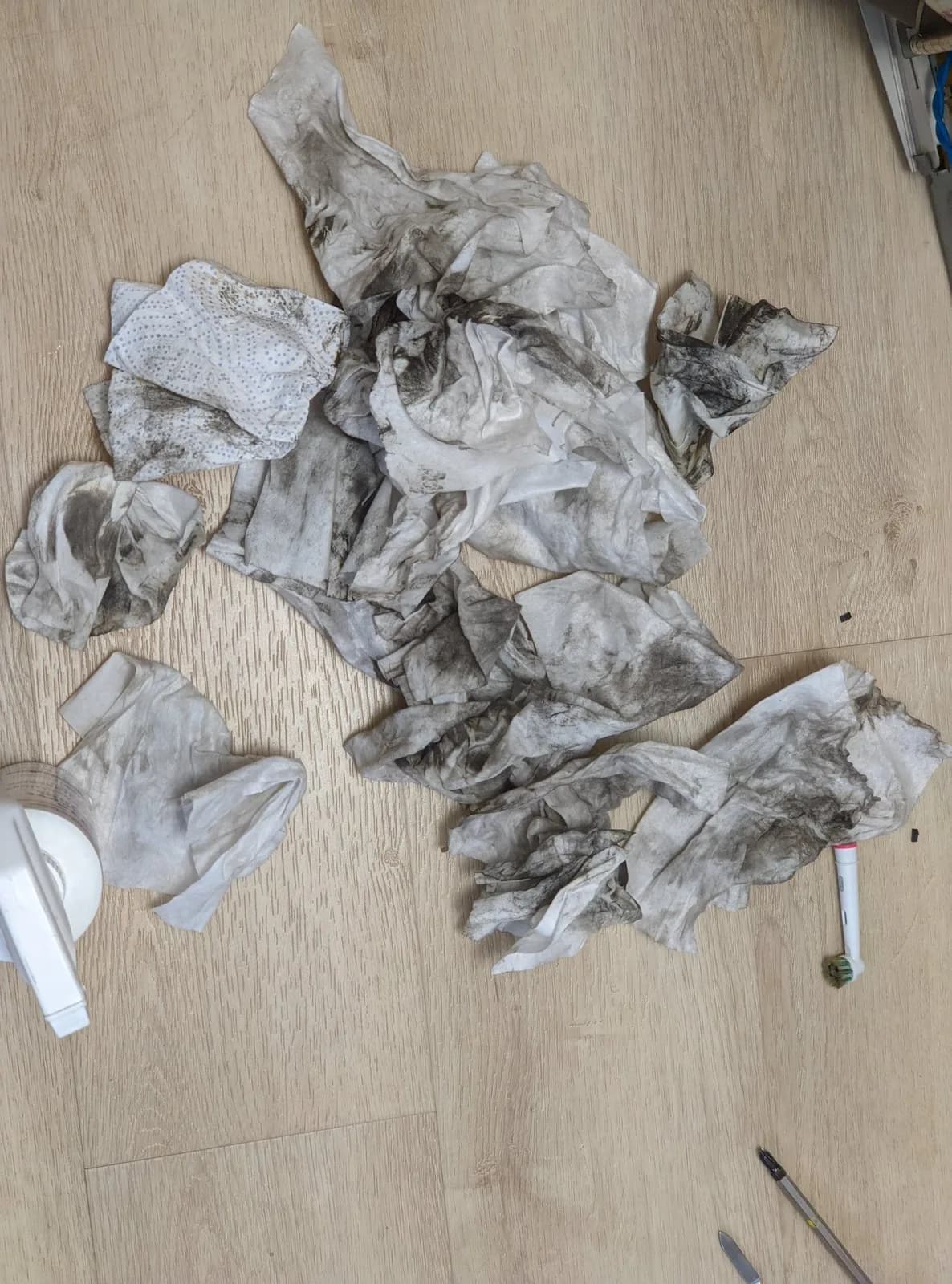 A pile of used cleaning wipes and paper towels stained grey and black with nicotine and grime on a wood floor, beside a white spray bottle of isopropyl alcohol, an electric-toothbrush head used for scrubbing, and a screwdriver at the edge of frame.