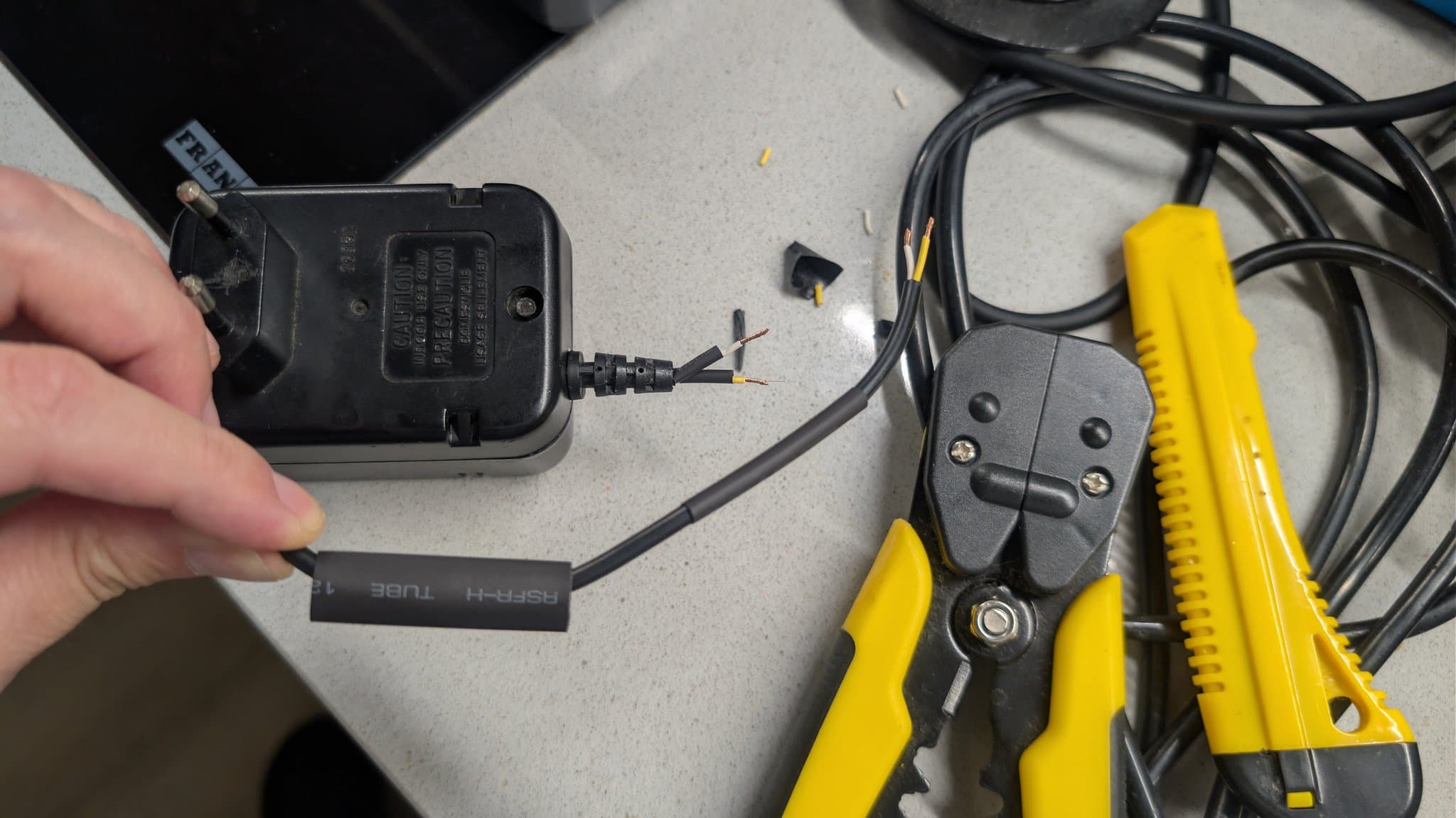 Hands holding stripped power connector with exposed wires next to crimper tool and heat shrink tubing during Game Gear power repair.