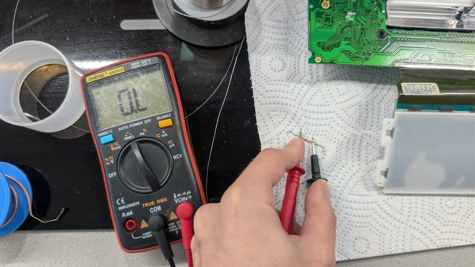 Digital multimeter displaying OL reading held over Game Gear PCB with green circuit board visible; fuse replacement repair in progress.