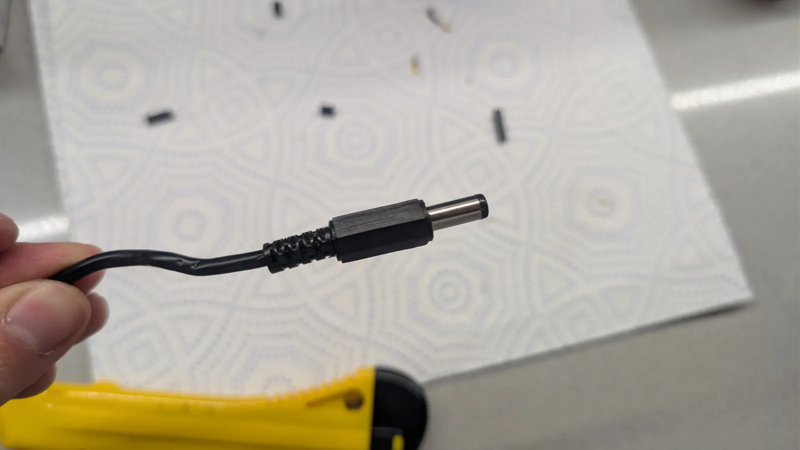 Close-up of barrel connector and black power cable for Sega Game Gear external PSU restoration against patterned white background.
