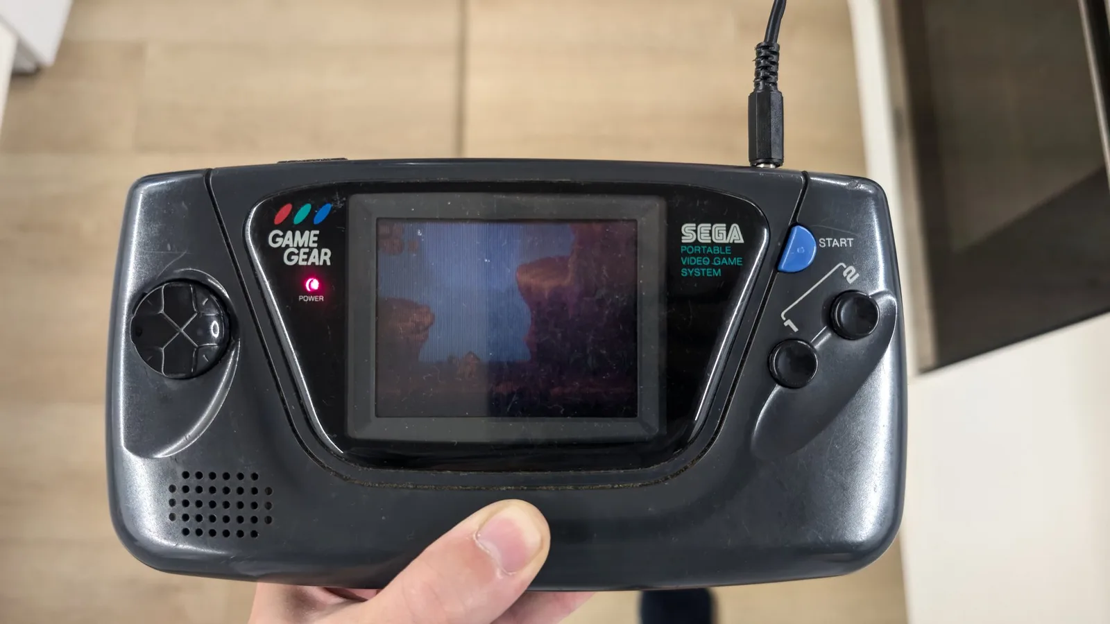 Sega Game Gear held in hand displaying repaired screen with underwater scene, power LED lit, external PSU restored.