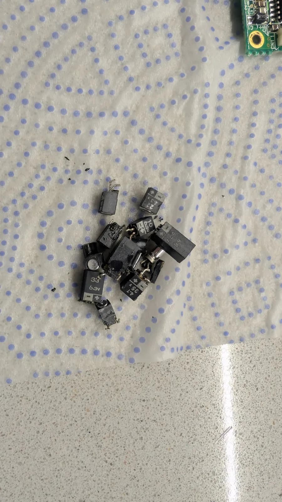 Pile of desoldered electrolytic capacitors from Sega Game Gear main board on dotted fabric, with circuit board visible top-right.