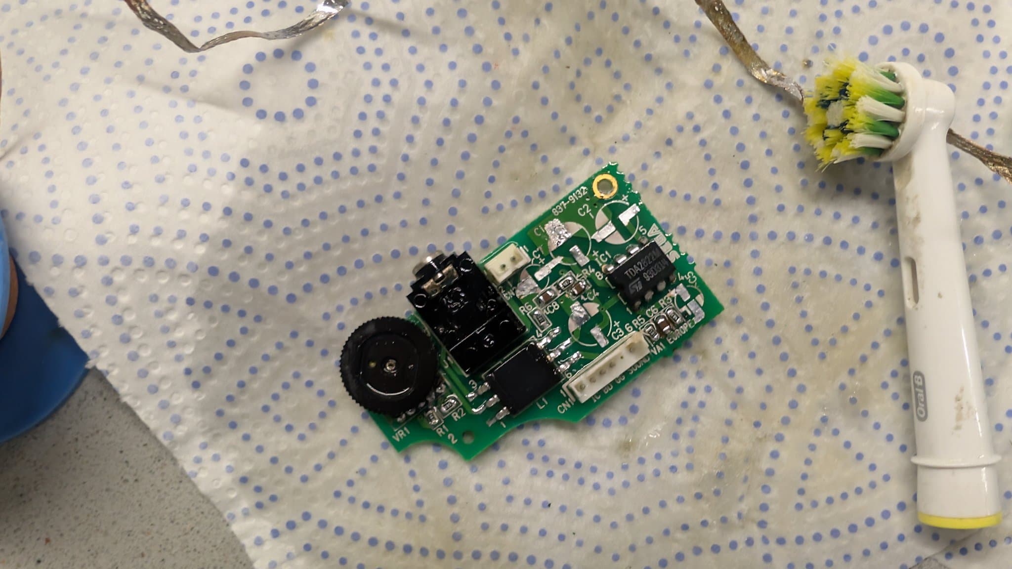 Game Gear audio/power board after ultrasonic cleaning, laid on dot matrix mat with soldering iron and yellow-flowered plant visible.