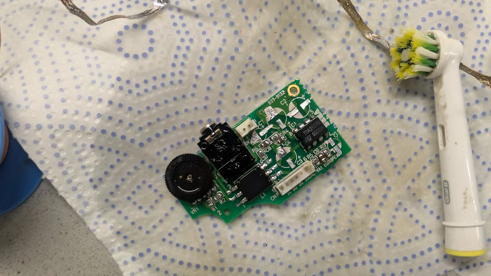 Game Gear audio/power board after ultrasonic cleaning, laid on dot matrix mat with soldering iron and yellow-flowered plant visible.