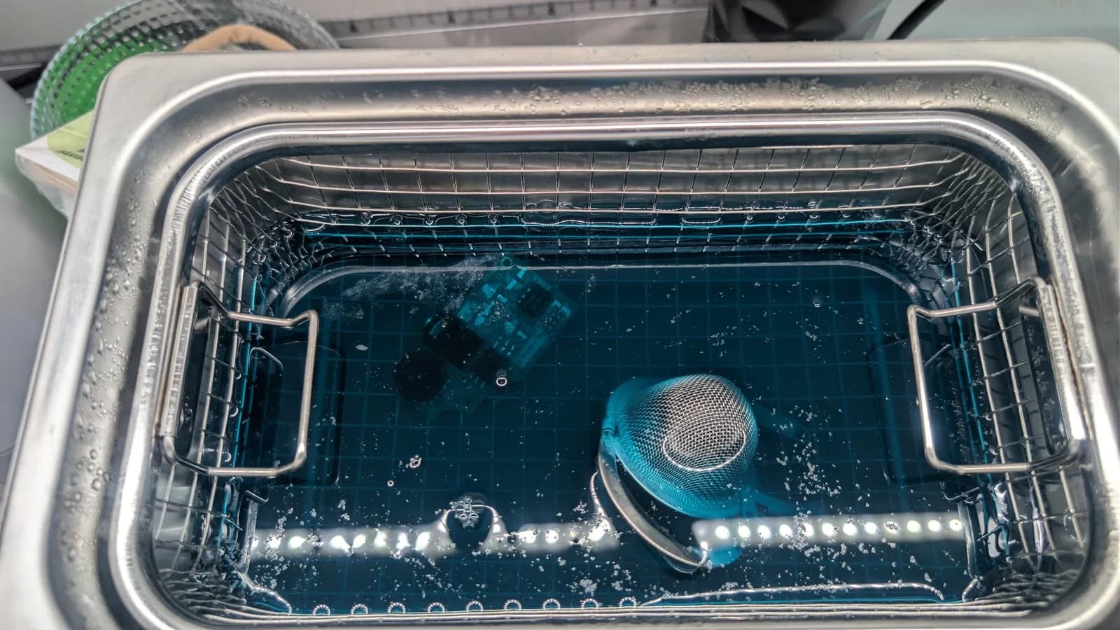 Sega Game Gear audio and power boards in ultrasonic cleaner with blue solution and mesh basket visible.