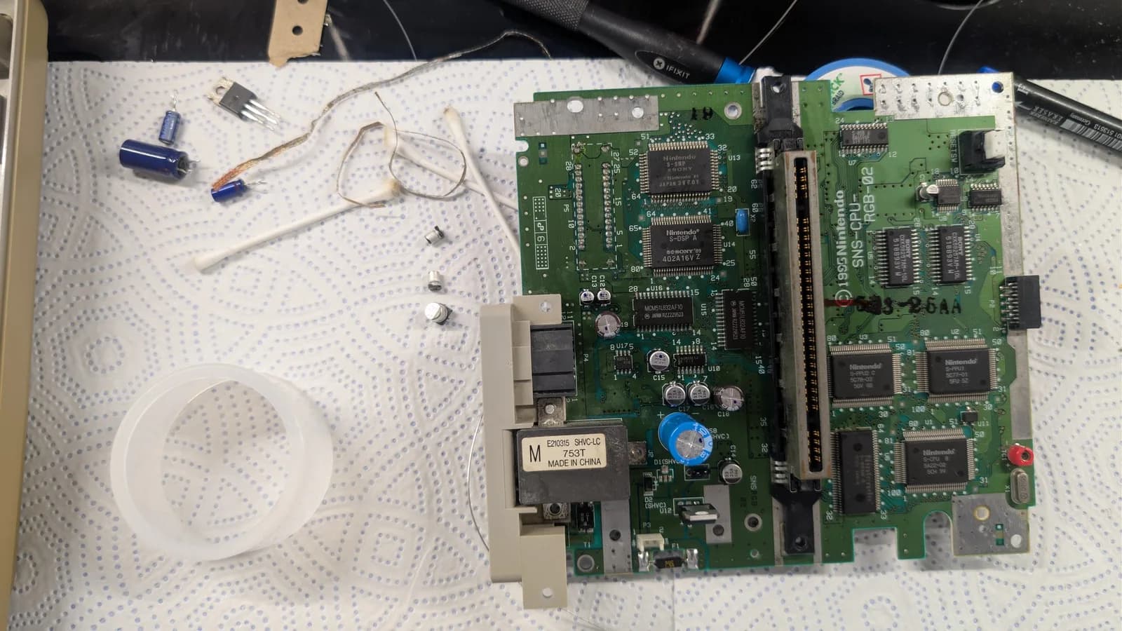 Super Famicom motherboard mid-repair: recapped with new capacitors, voltage regulator replaced, some SMD components remaining; soldering iron and loose components visible on workbench.