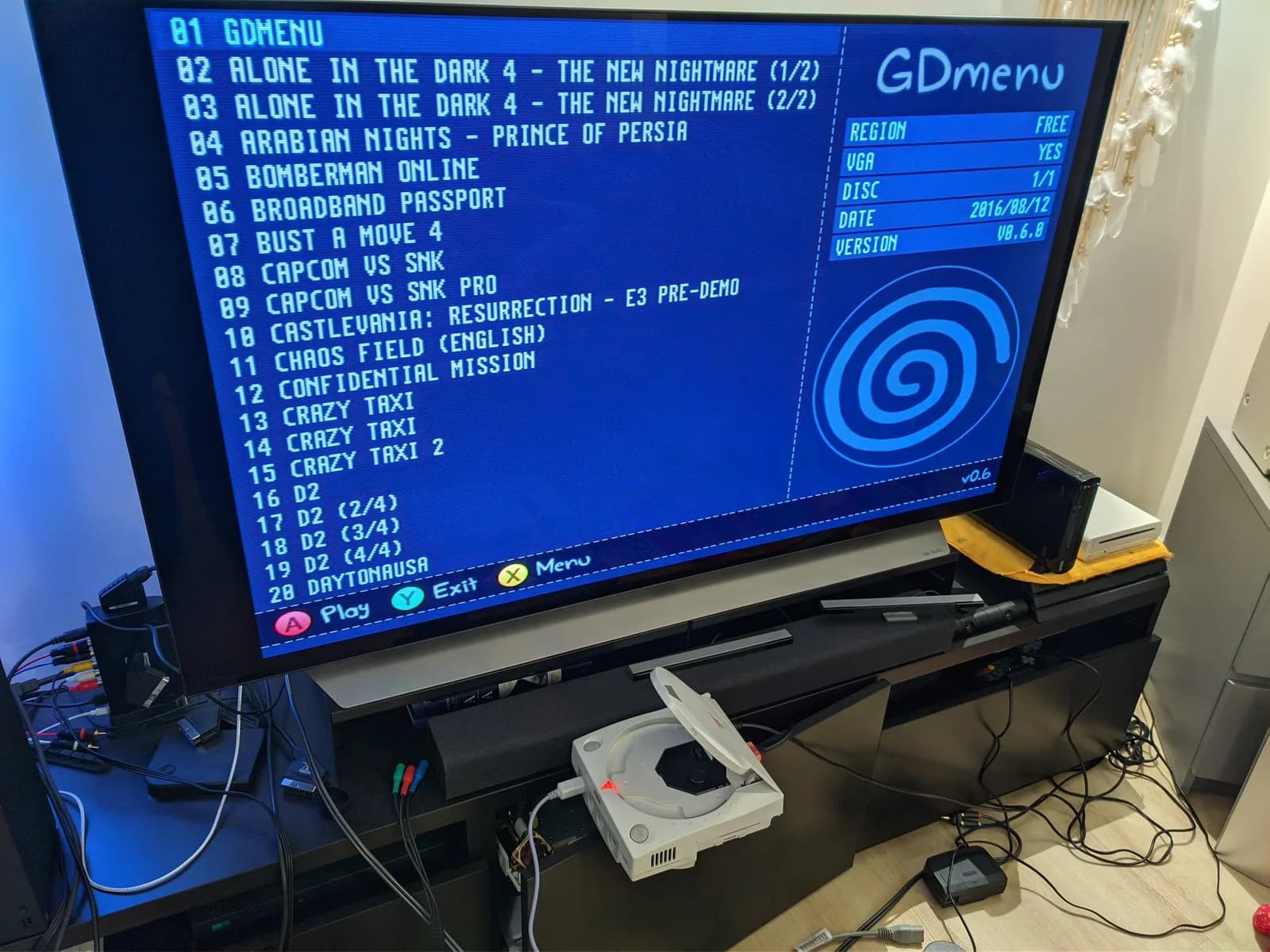 A large TV shows the GDmenu game-selection screen (Dreamcast swirl logo, Region Free, VGA Yes, v0.6.0); below it the white Sega Dreamcast VA0, running the installed GDEMU, is propped on the half-open door of the TV stand.
