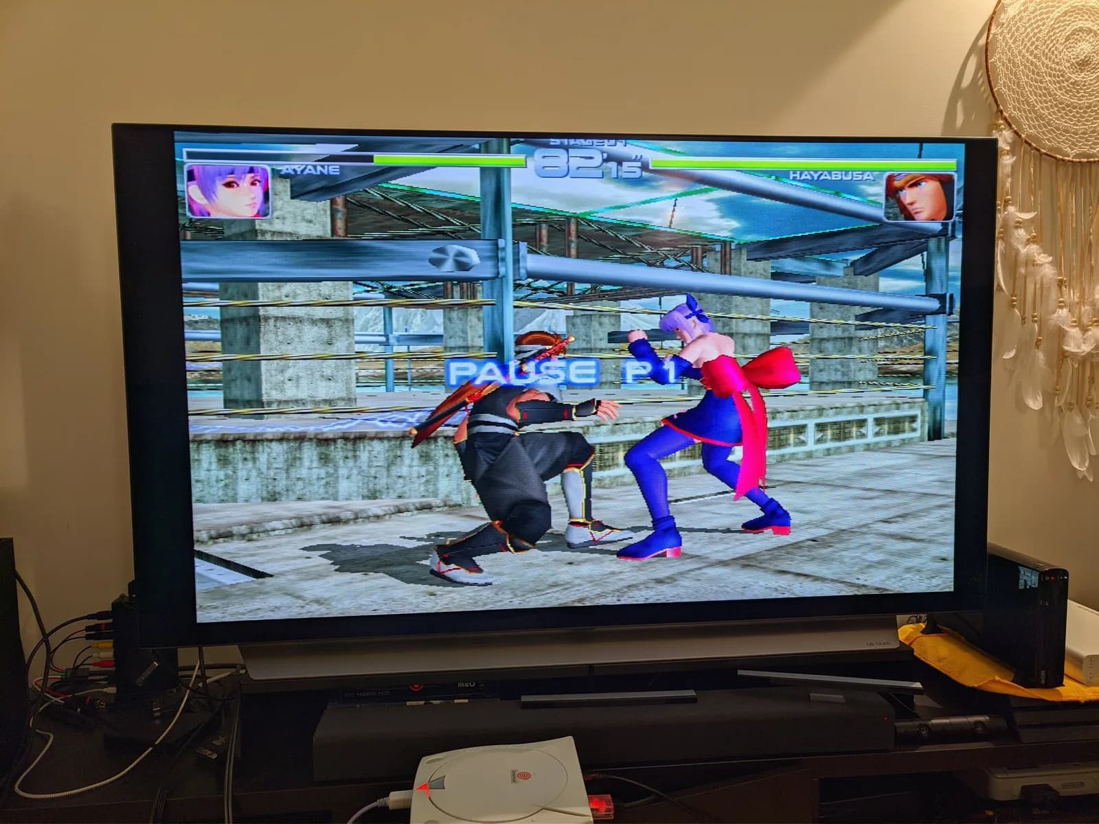 A flatscreen TV shows Dead or Alive 2 (Ayane versus Hayabusa) paused; the white Sega Dreamcast VA0, running it through the installed GDEMU, sits on the stand below.