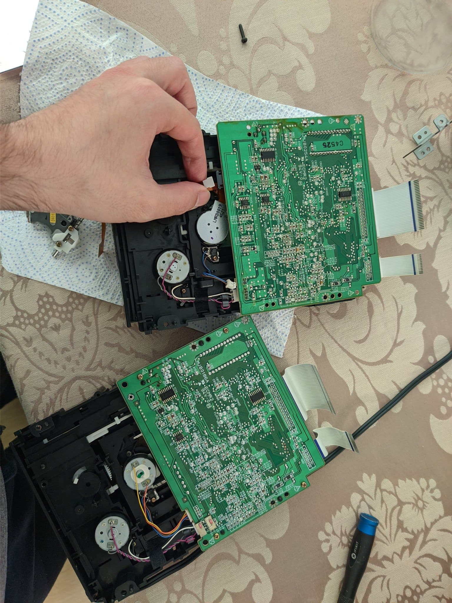 Technician's hand holding black plastic component above disassembled Panasonic FZ-1 3DO console showing green PCBs, internal mechanisms, and ribbon cables during laser replacement repair.