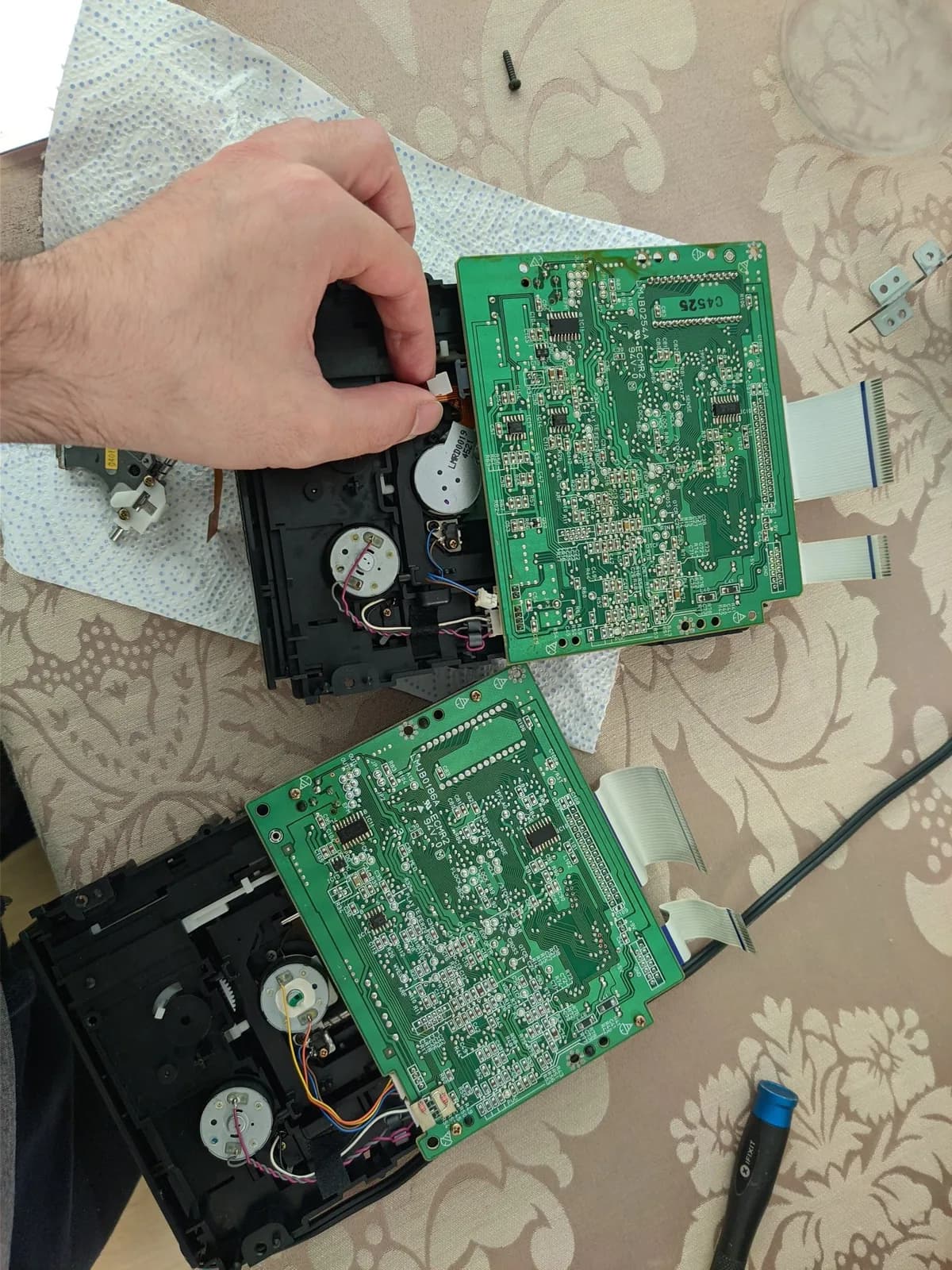 Technician's hand holding black plastic component above disassembled Panasonic FZ-1 3DO console showing green PCBs, internal mechanisms, and ribbon cables during laser replacement repair.