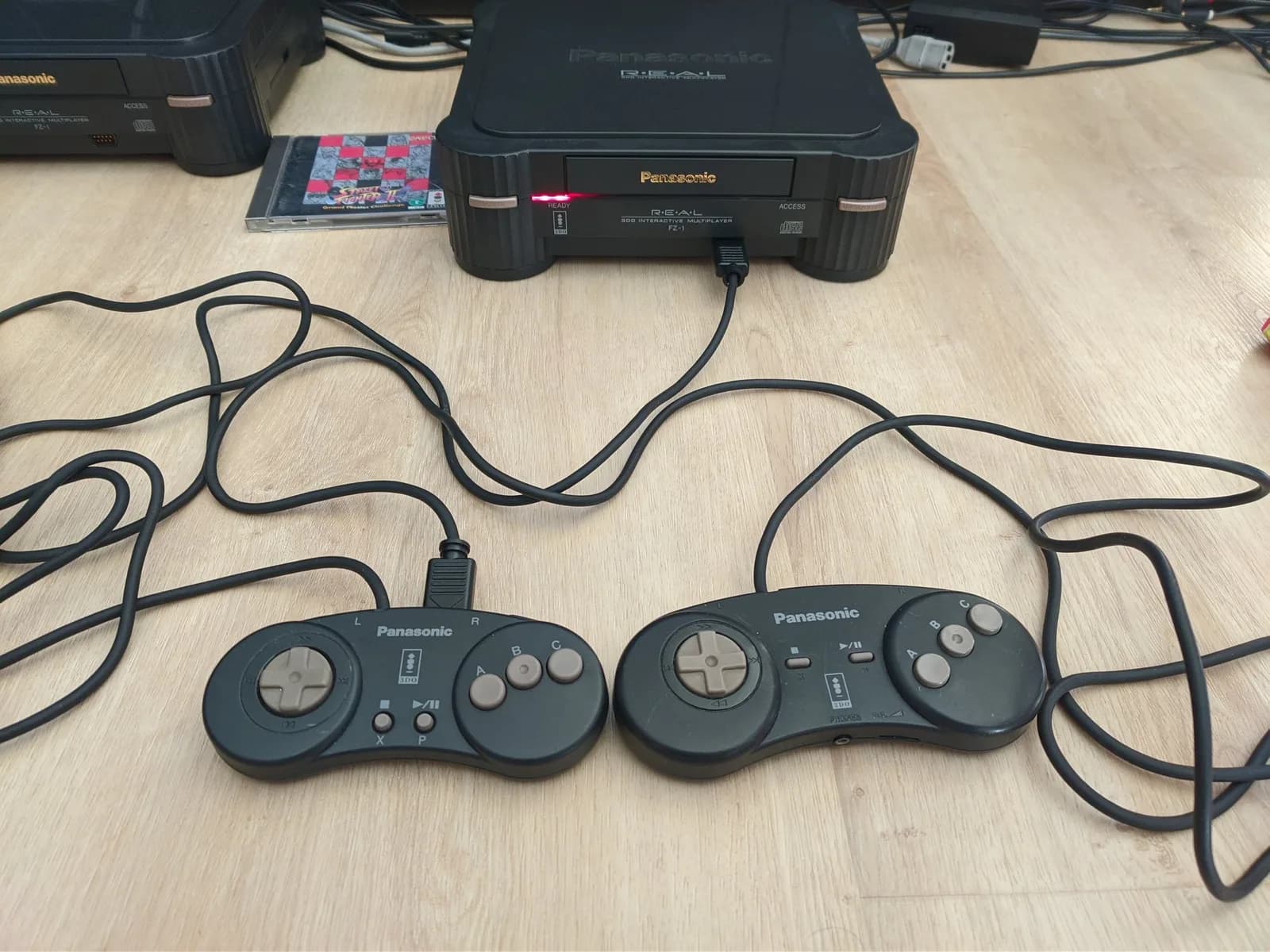 Panasonic 3DO FZ-1 console with two daisy-chained controllers connected via coiled cables, displaying S-video mod setup on wooden surface.