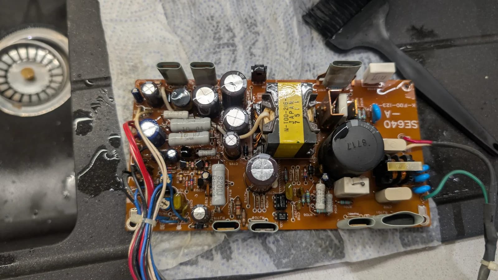 The Sharp X68000 PSU board removed from its housing and soaked in white vinegar to neutralize the alkaline electrolyte, its brown PCB with multiple electrolytic capacitors, the transformer and residue from the failed caps.