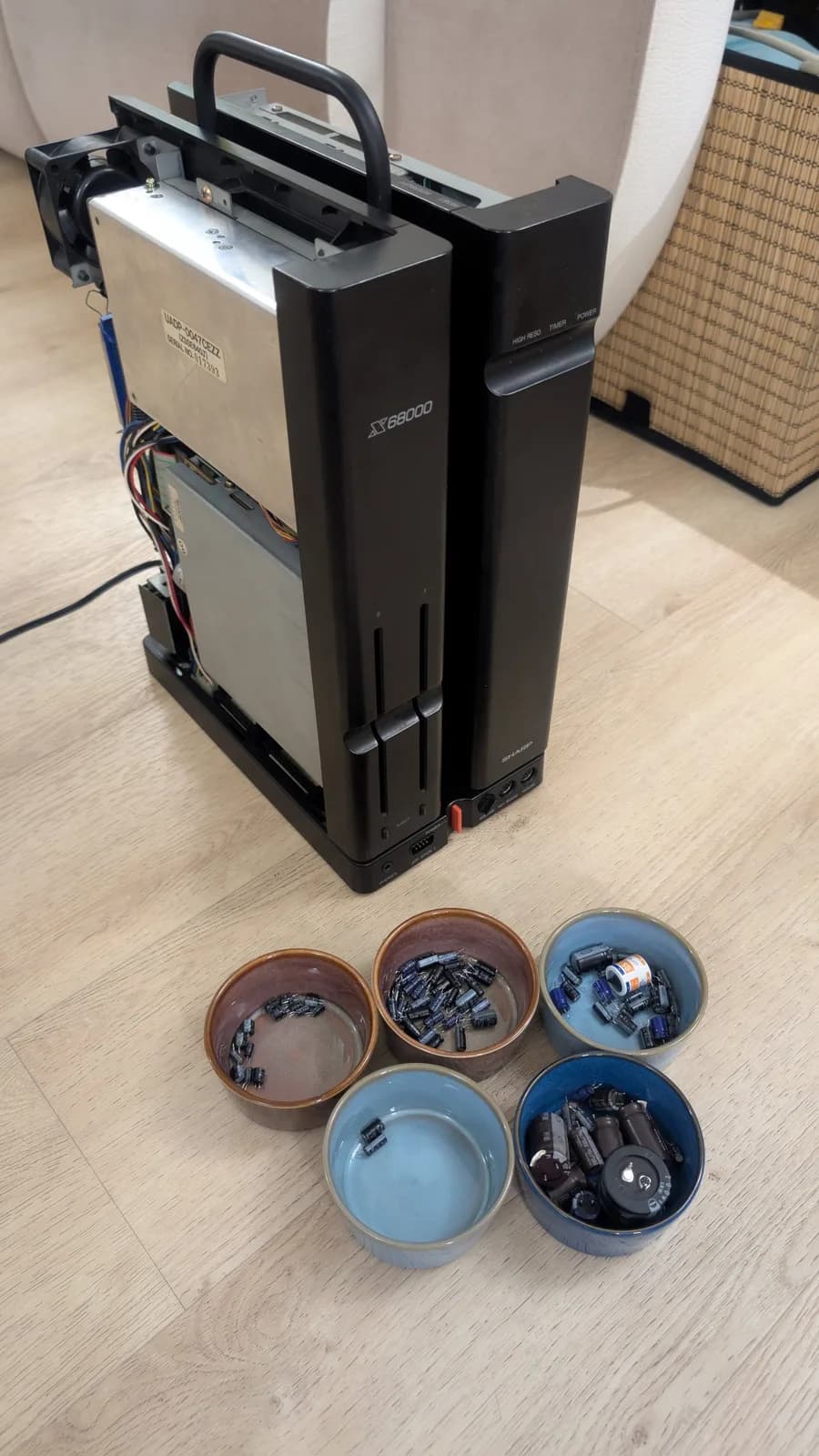 The old capacitors from each board sorted into their own bowls on the floor along with the old battery, the Sharp X68000 main unit with its carry handle in the background, ready for reassembly.