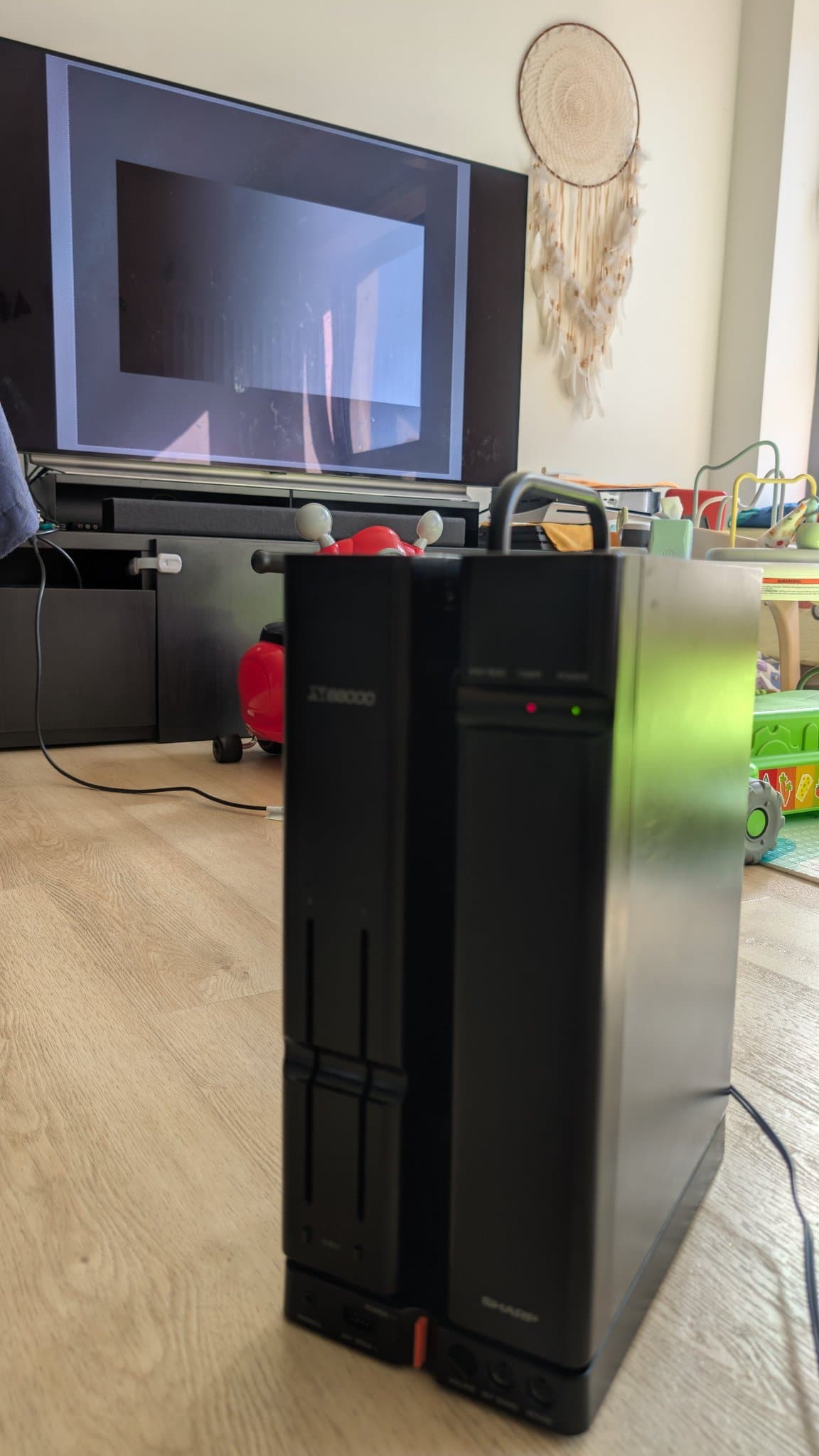Black Sharp X68000 tower on wooden floor displaying no video output on connected monitor despite power-on state.