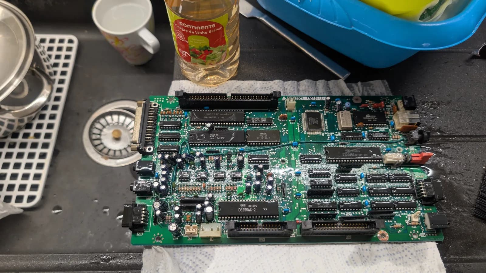 The Sharp X68000 lower board being doused in white vinegar to neutralize the leaked electrolyte, a brush and the white vinegar bottle alongside.