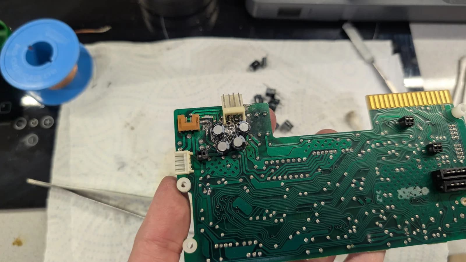 The recapped FDD controller PCB held in hand, its new capacitors fitted, the old capacitors and desoldering braid on the paper towel in the background.