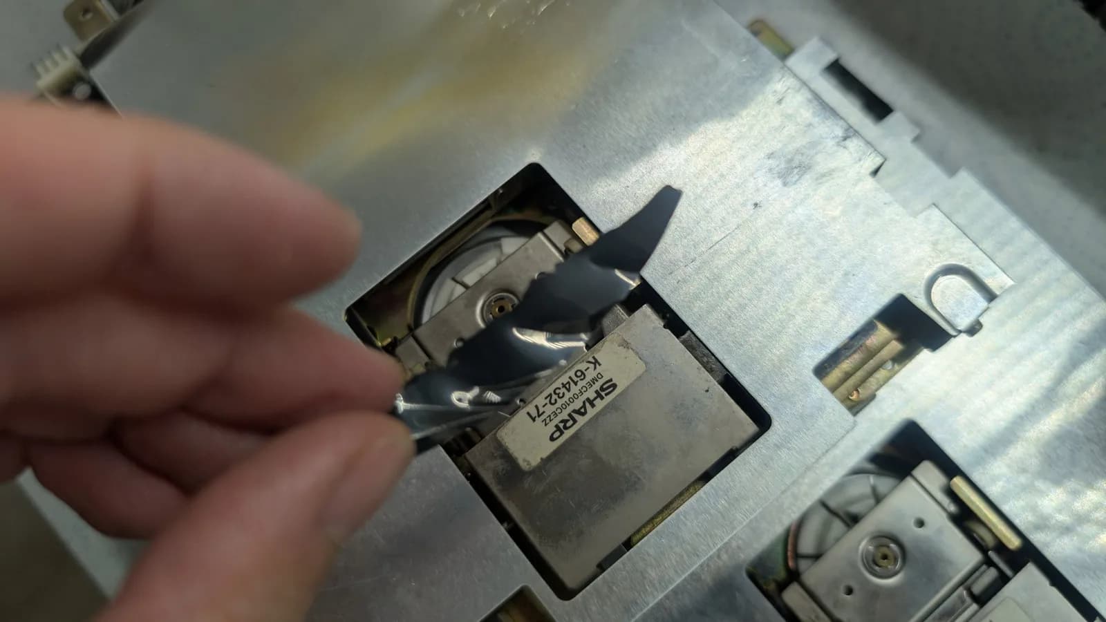 A close-up of the torn magnetic-disk fragment held above the floppy drive, the drive beneath in its metal housing with the spindle mechanism showing.