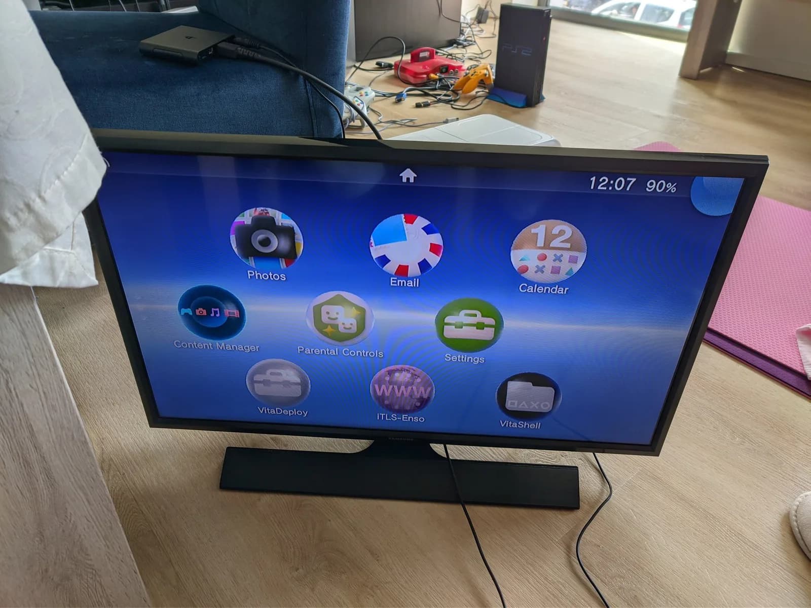 Sony Vita TV displaying home menu with apps, mounted on black stand on wooden desk with gaming equipment visible behind.