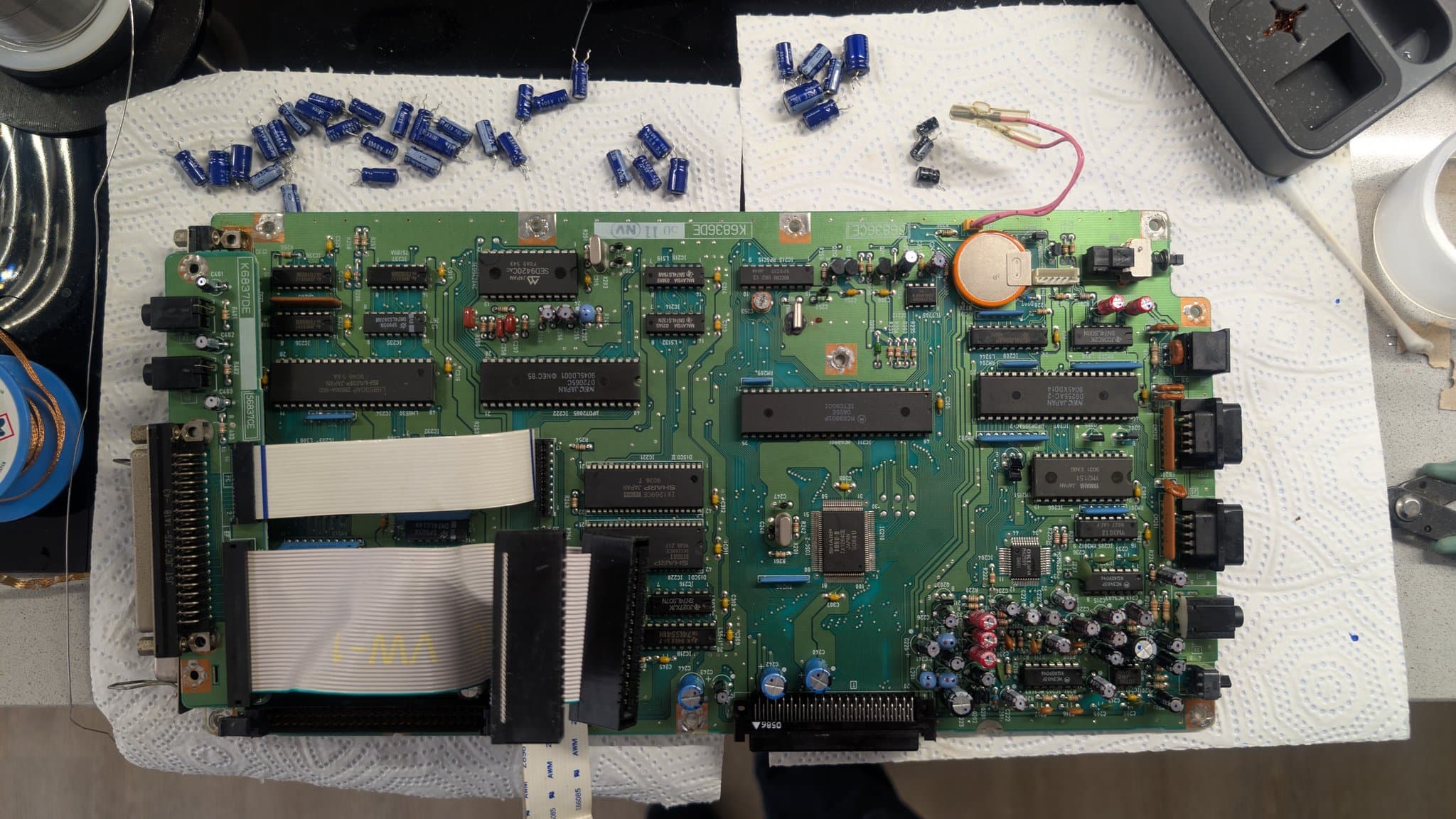 The Sharp X68000 Pro II control board mid-recap on paper towels, the new capacitors already installed on the board and the old blue capacitors set above it, two ribbon cables detached and the orange battery still soldered in place.