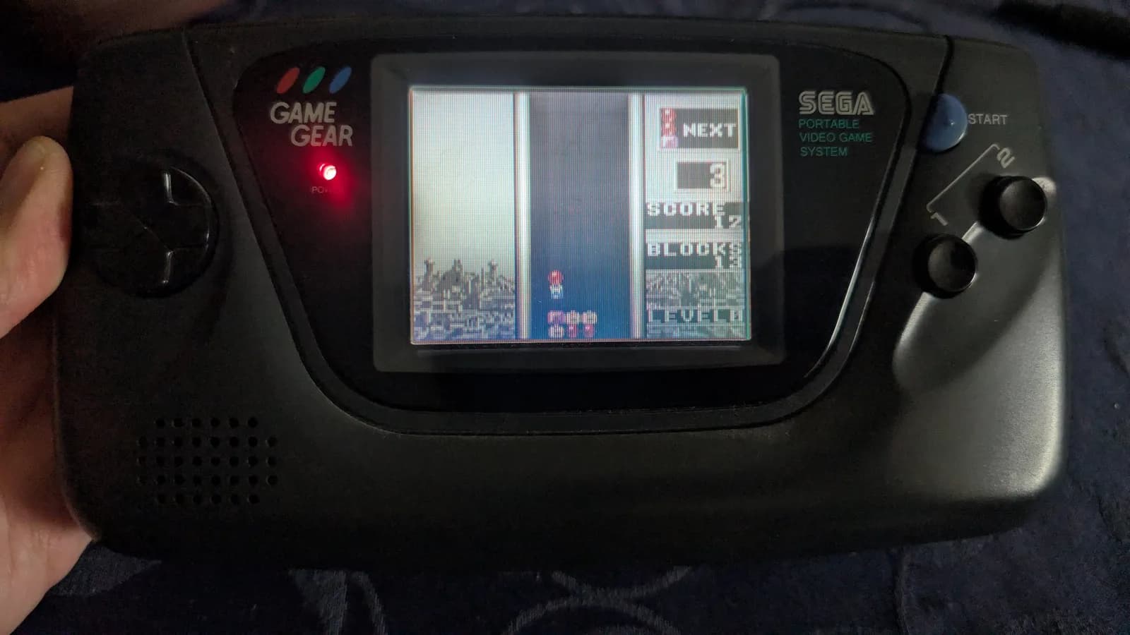 Sega Game Gear displaying menu screen with three game options visible, held in hands during ceramic capacitor replacement repair.