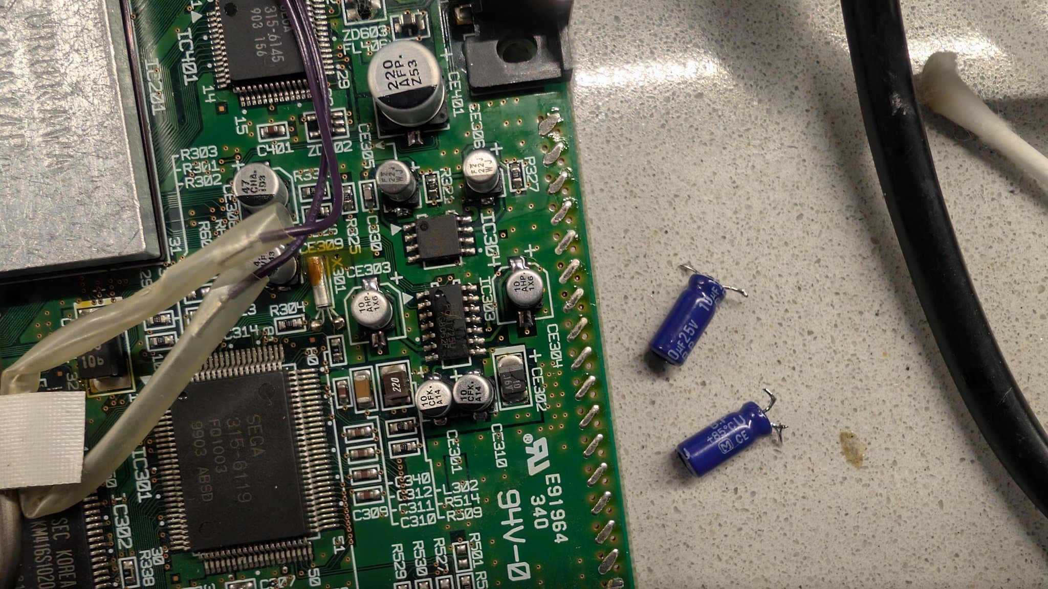 Two small blue radial capacitors on the bench beside a Dreamcast board, the stopgap radial parts used while the proper bi-polar capacitors were out of stock.