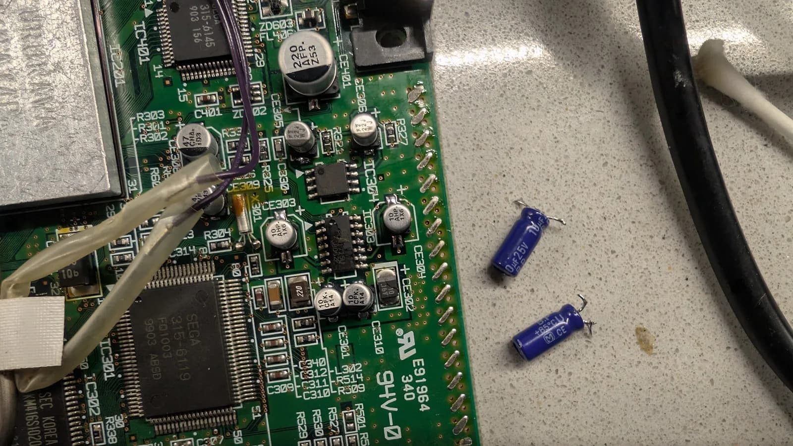 Two small blue radial capacitors on the bench beside a Dreamcast board, the stopgap radial parts used while the proper bi-polar capacitors were out of stock.