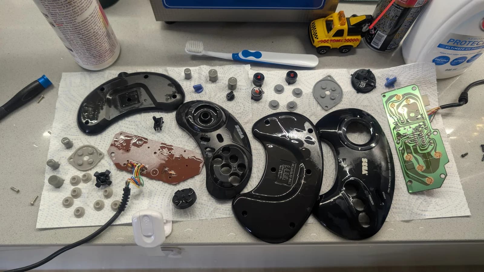 Disassembled Mega Drive controllers on white mat showing black plastic shells, circuit boards, button mechanisms, springs, and small components during deep cleaning restoration.