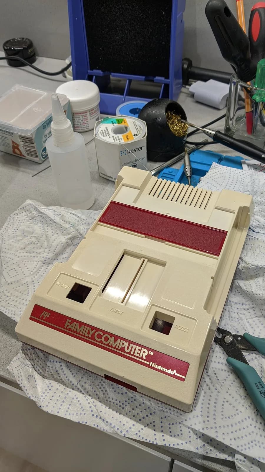 Repaired Famicom with composite mod board installed, displayed on workbench with soldering iron, flux, and cleaning supplies nearby.