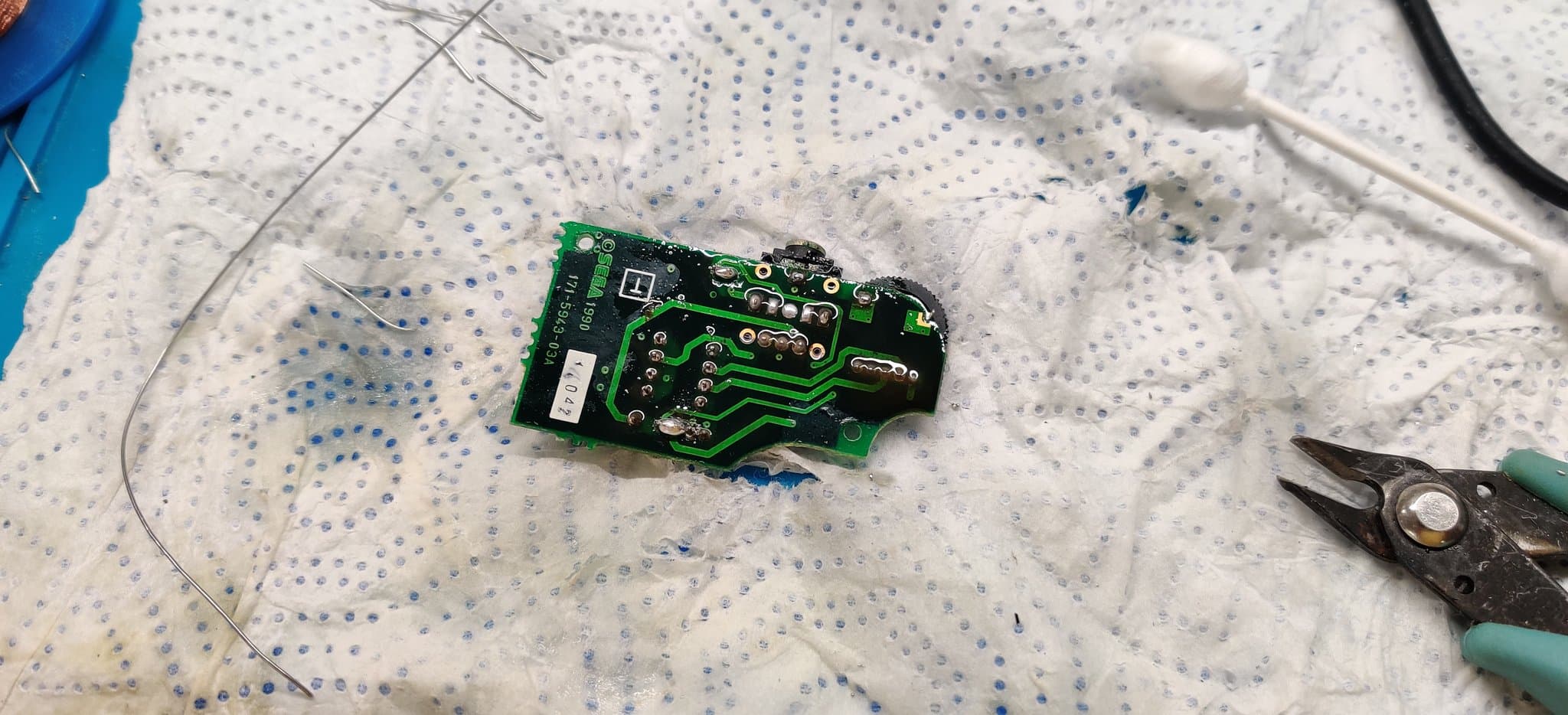 A Game Gear sound board after its solder was reflowed to clean off electrolyte contamination from the old capacitors, the board still wet with flux, with a pair of pliers and a length of solder wire alongside.