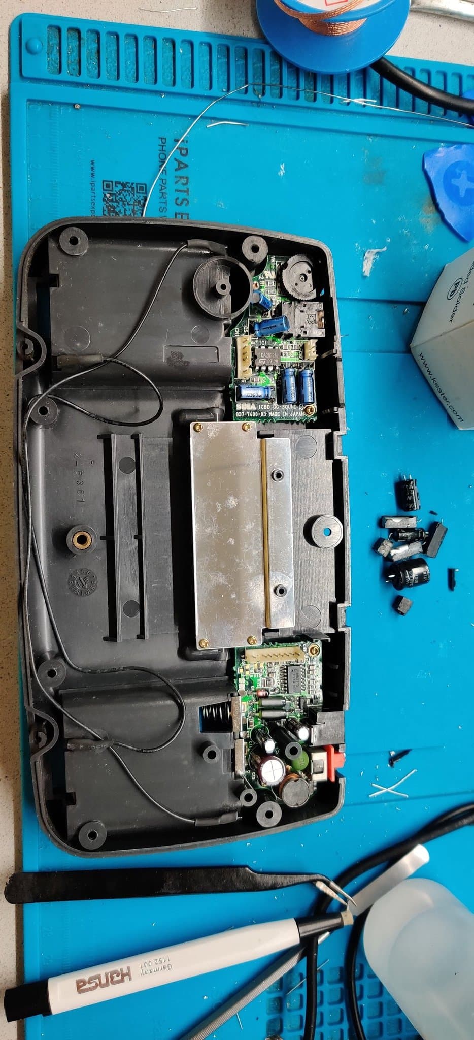 The rear half of one Game Gear's shell with its sound and power boards refitted after recapping, the removed old capacitors beside the shell and a pair of tweezers, a fiberglass scratch pen and a metal spudger on the mat.