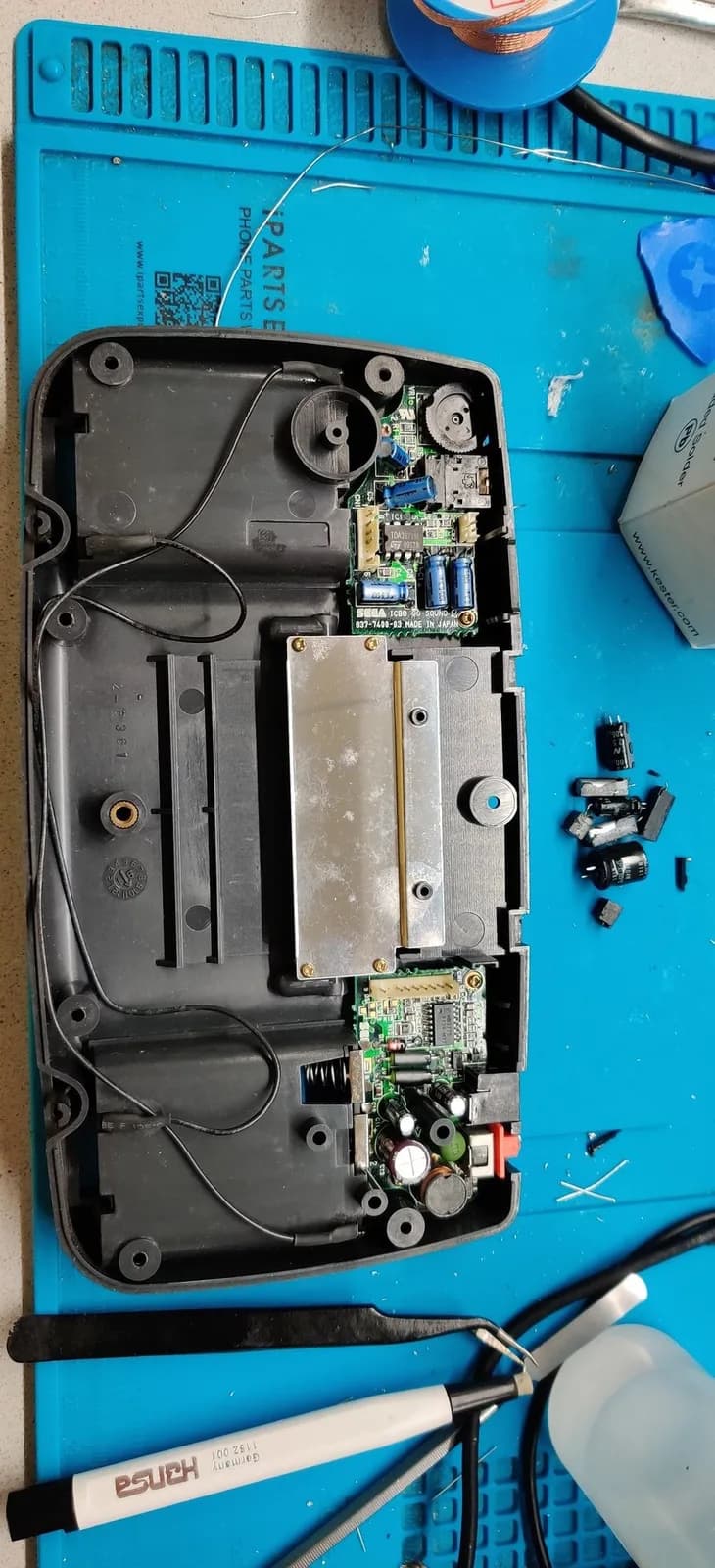 The rear half of one Game Gear's shell with its sound and power boards refitted after recapping, the removed old capacitors beside the shell and a pair of tweezers, a fiberglass scratch pen and a metal spudger on the mat.