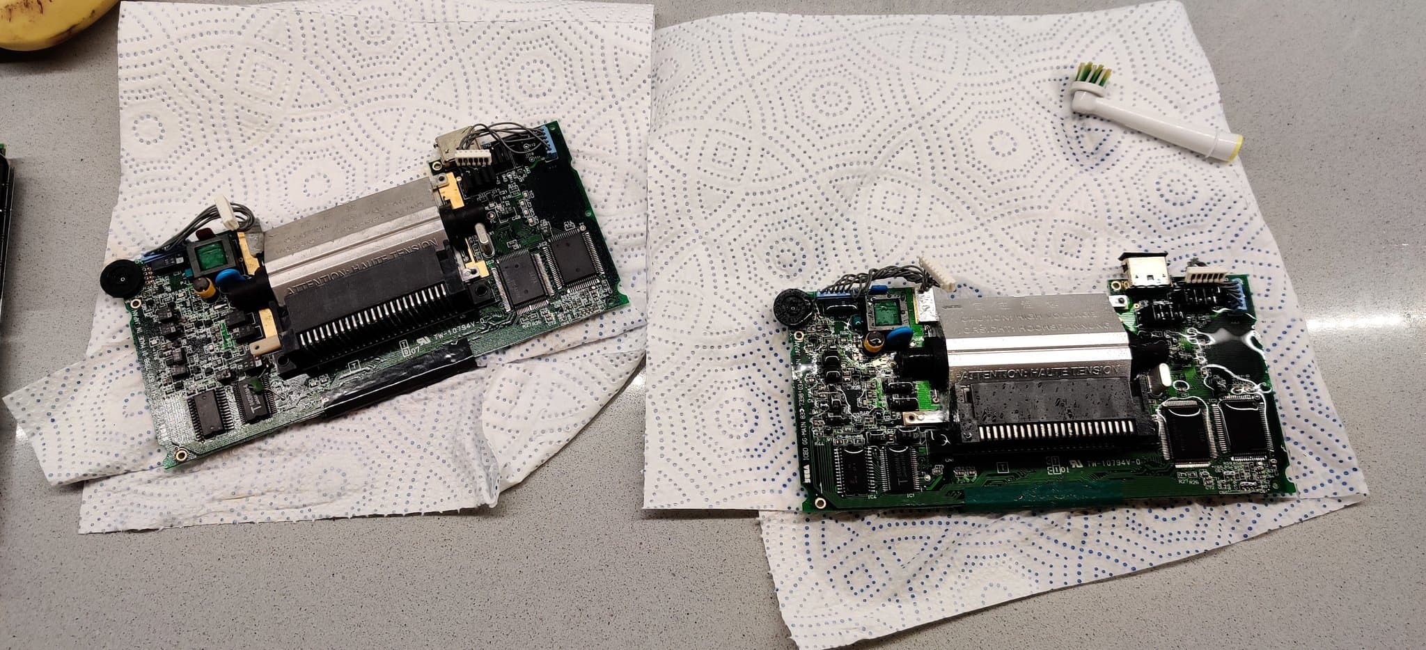 Both Game Gear mainboards on the mat being cleaned with white vinegar to neutralize the alkaline electrolyte, the board on the right still drenched and gleaming with pooled vinegar.